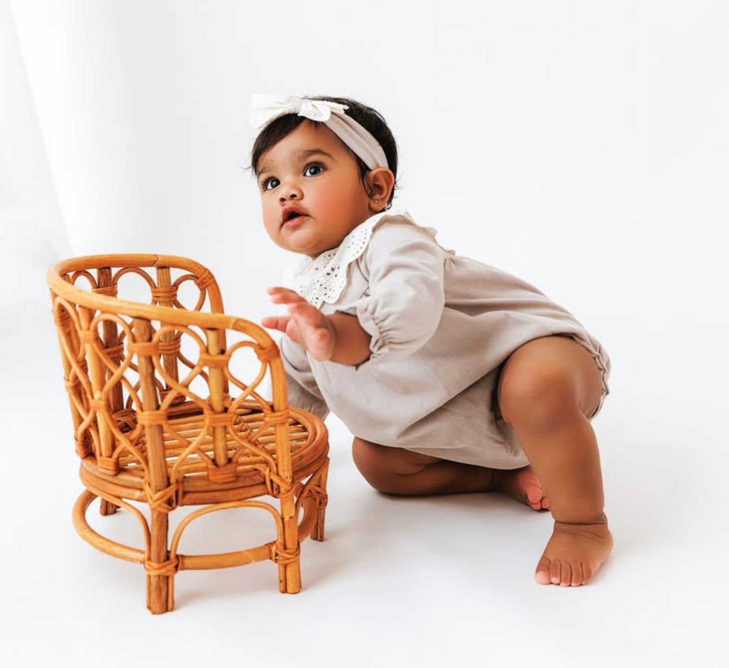 A baby in white clothing crawls and plays with a small wicker rattan chair against a white background.