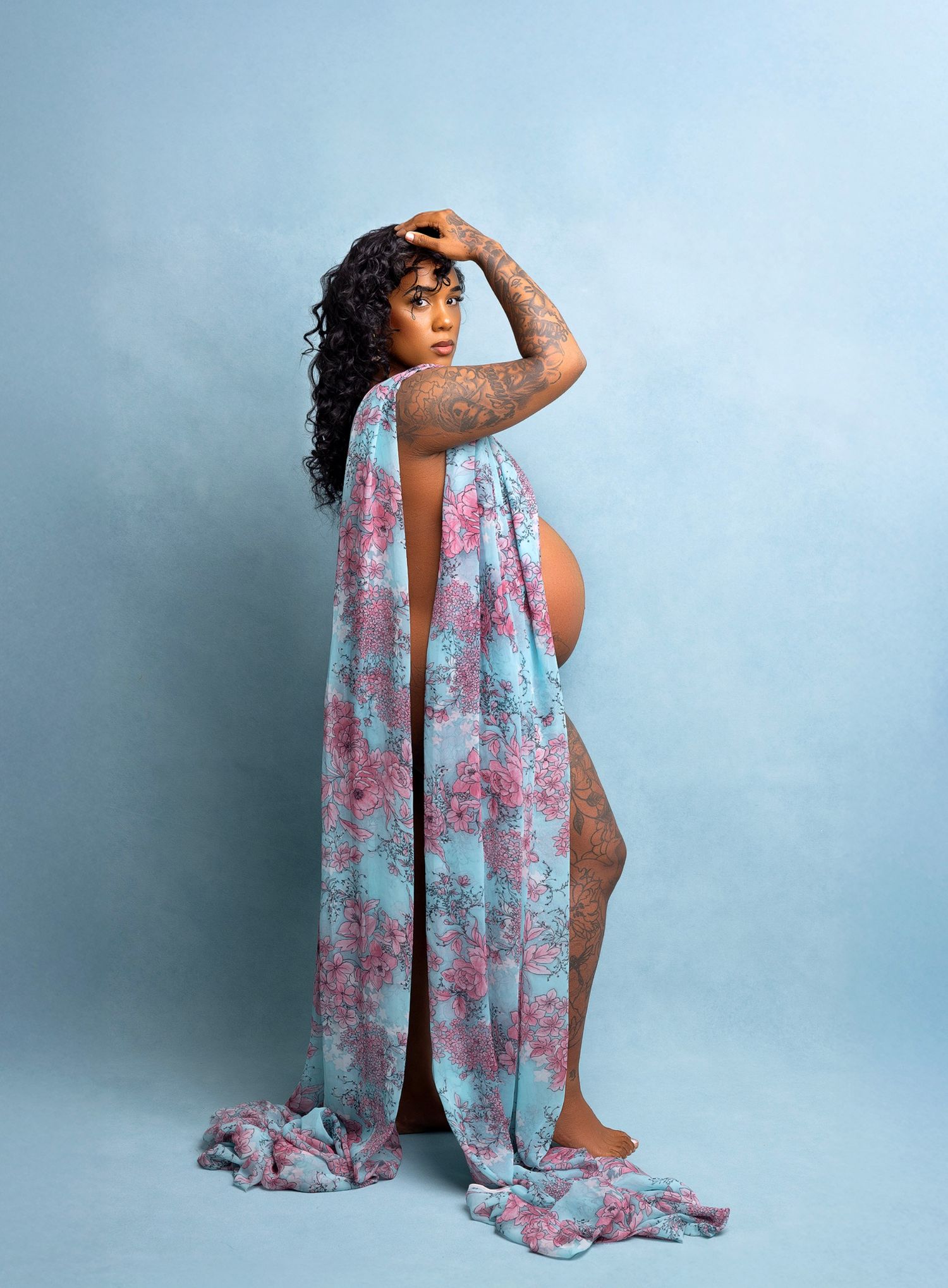 Artistic nude figure poses gracefully with flowing pink and blue floral fabric against light blue studio backdrop.