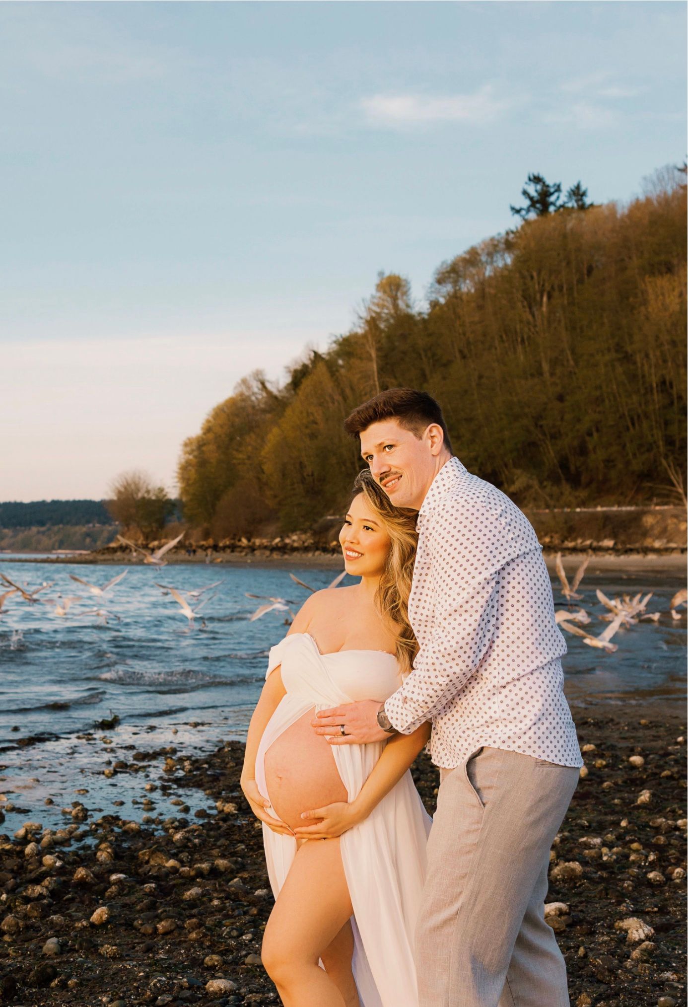 Pregnant couple posing on rocky beach at sunset, woman in white maternity dress, man embracing her baby bump.