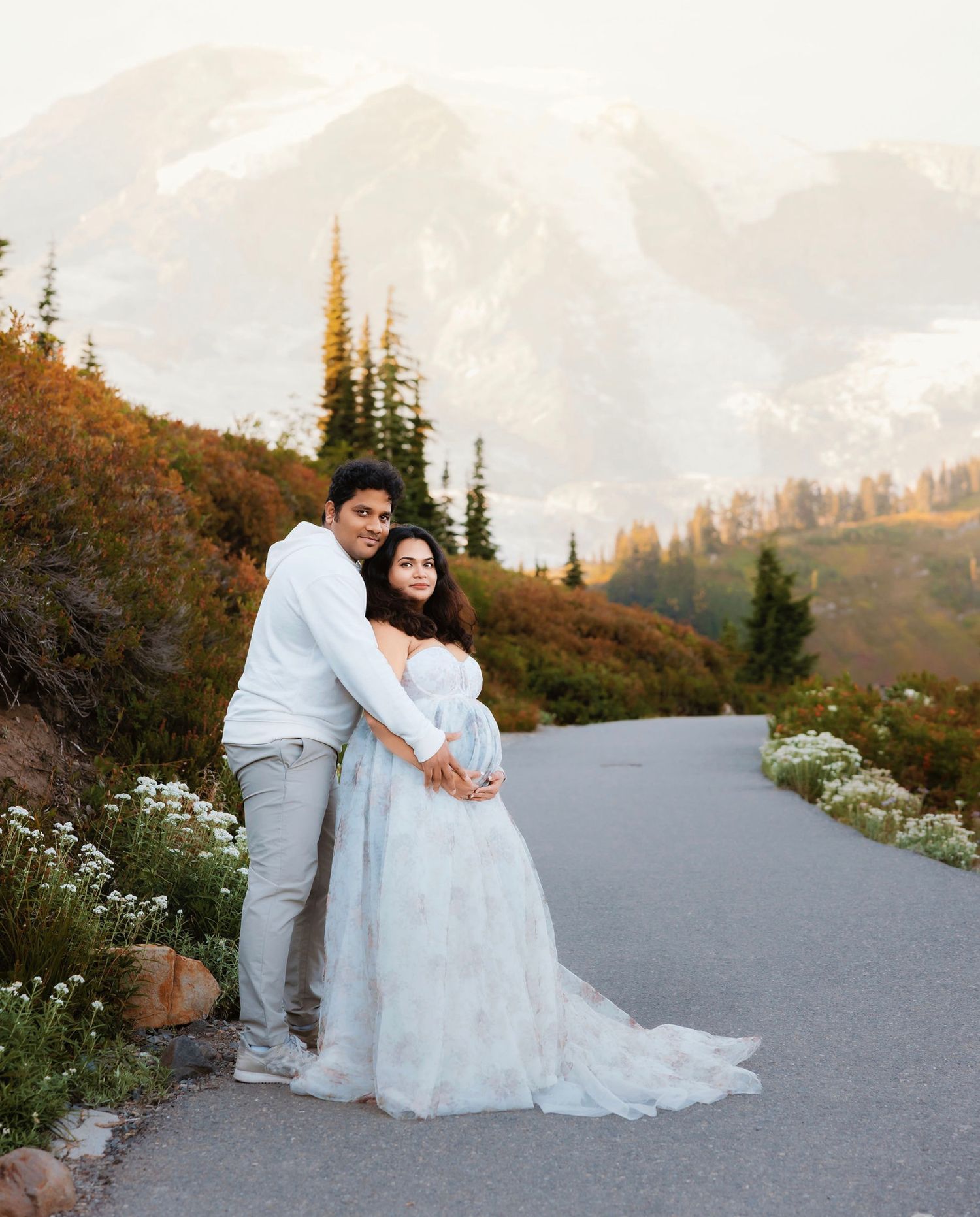 Couple in wedding attire embracing on a mountain road during sunset with evergreen trees in the background.