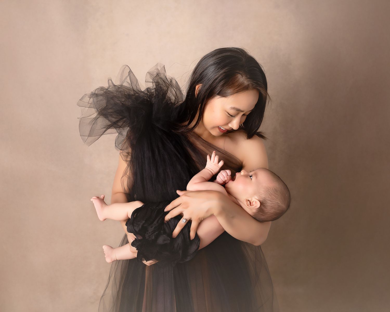 A mother in a black tulle dress tenderly holds her newborn baby against a warm beige studio backdrop.