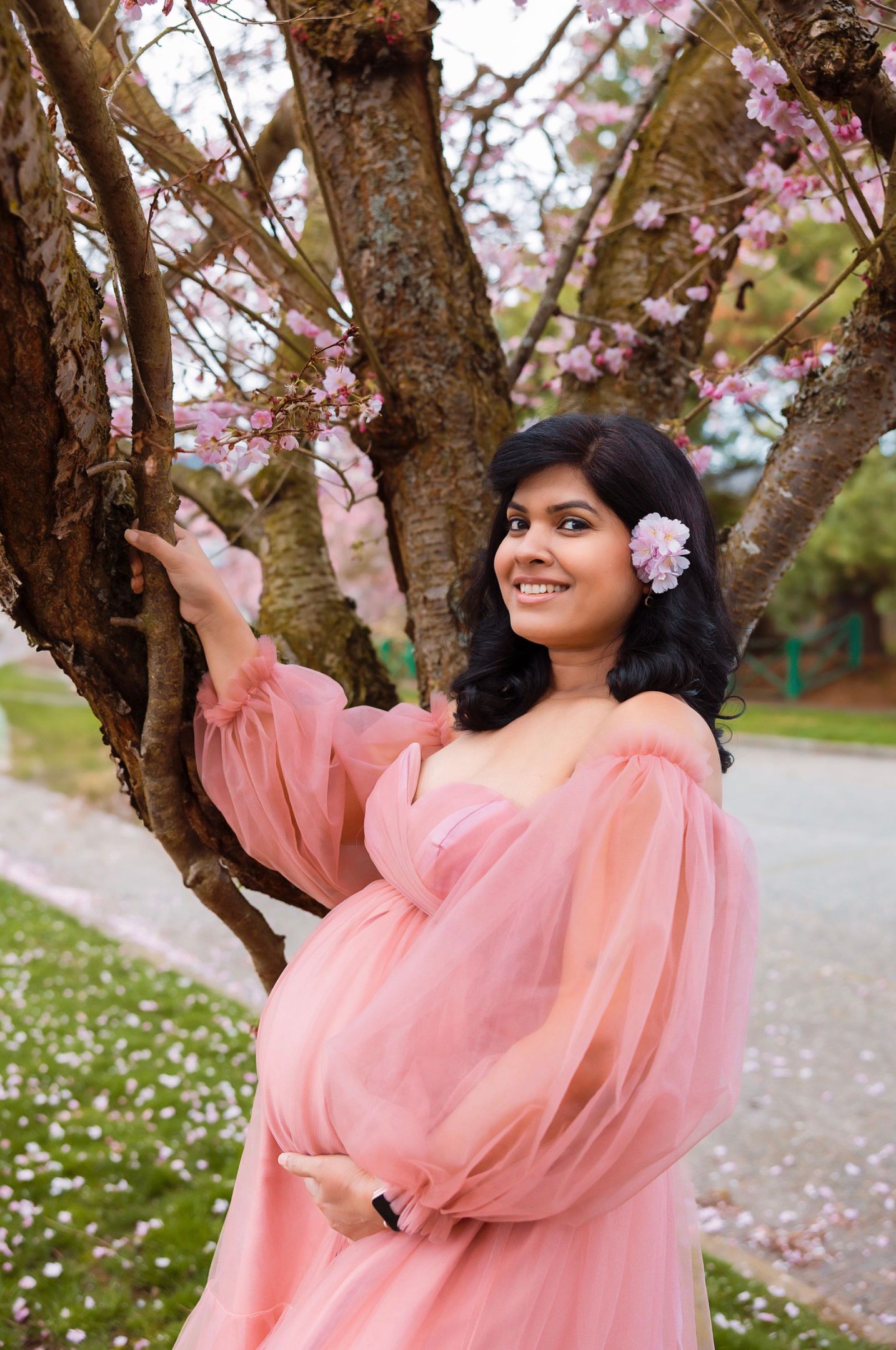 A woman in a flowing pink dress poses gracefully under blooming cherry blossom trees in spring.