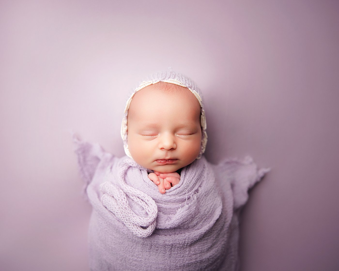 A sleeping newborn baby wrapped in soft purple fabric creates a dreamy portrait series against a matching background.