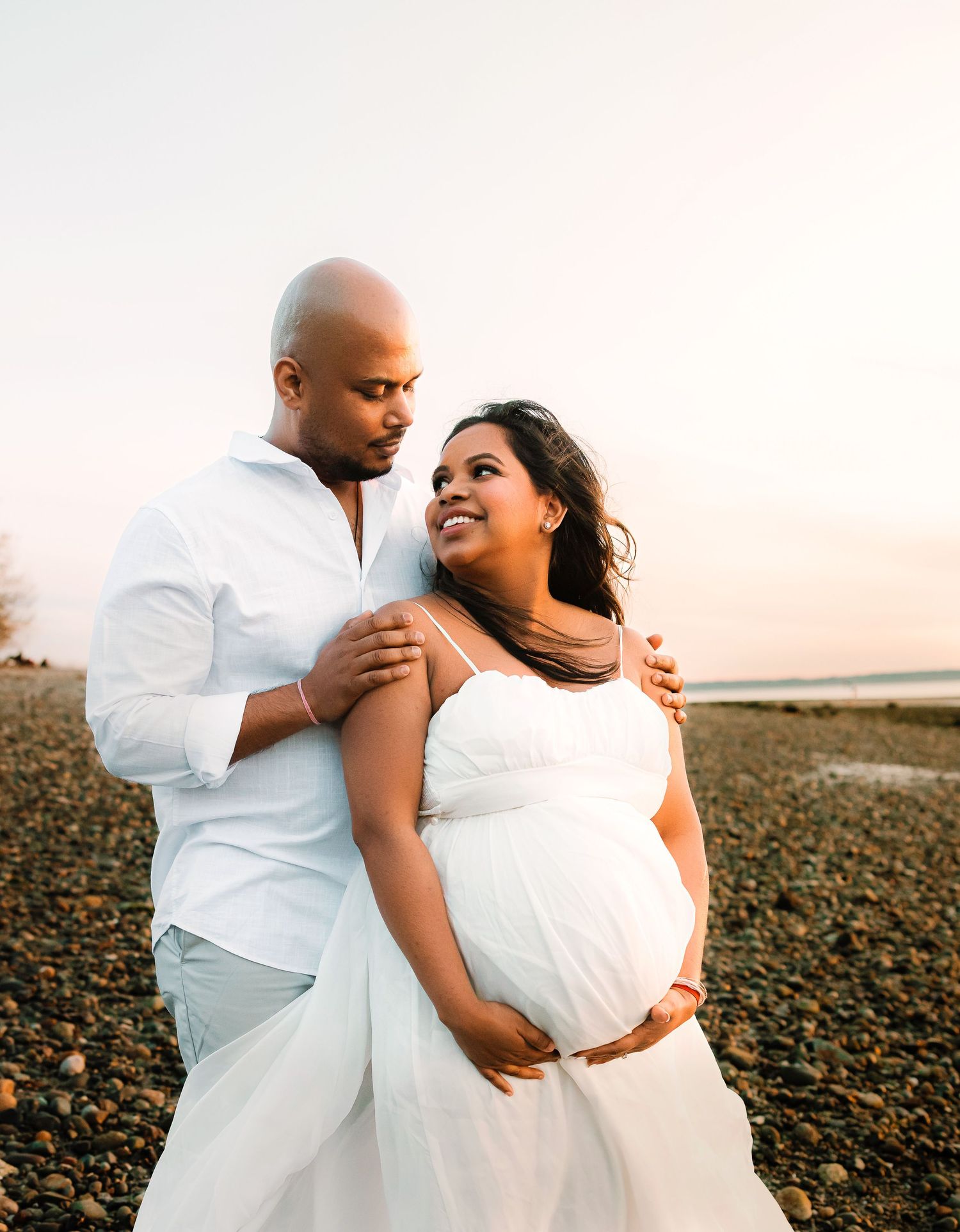 Pregnant couple in white outfits sharing a tender moment on a rocky beach at golden hour sunset.