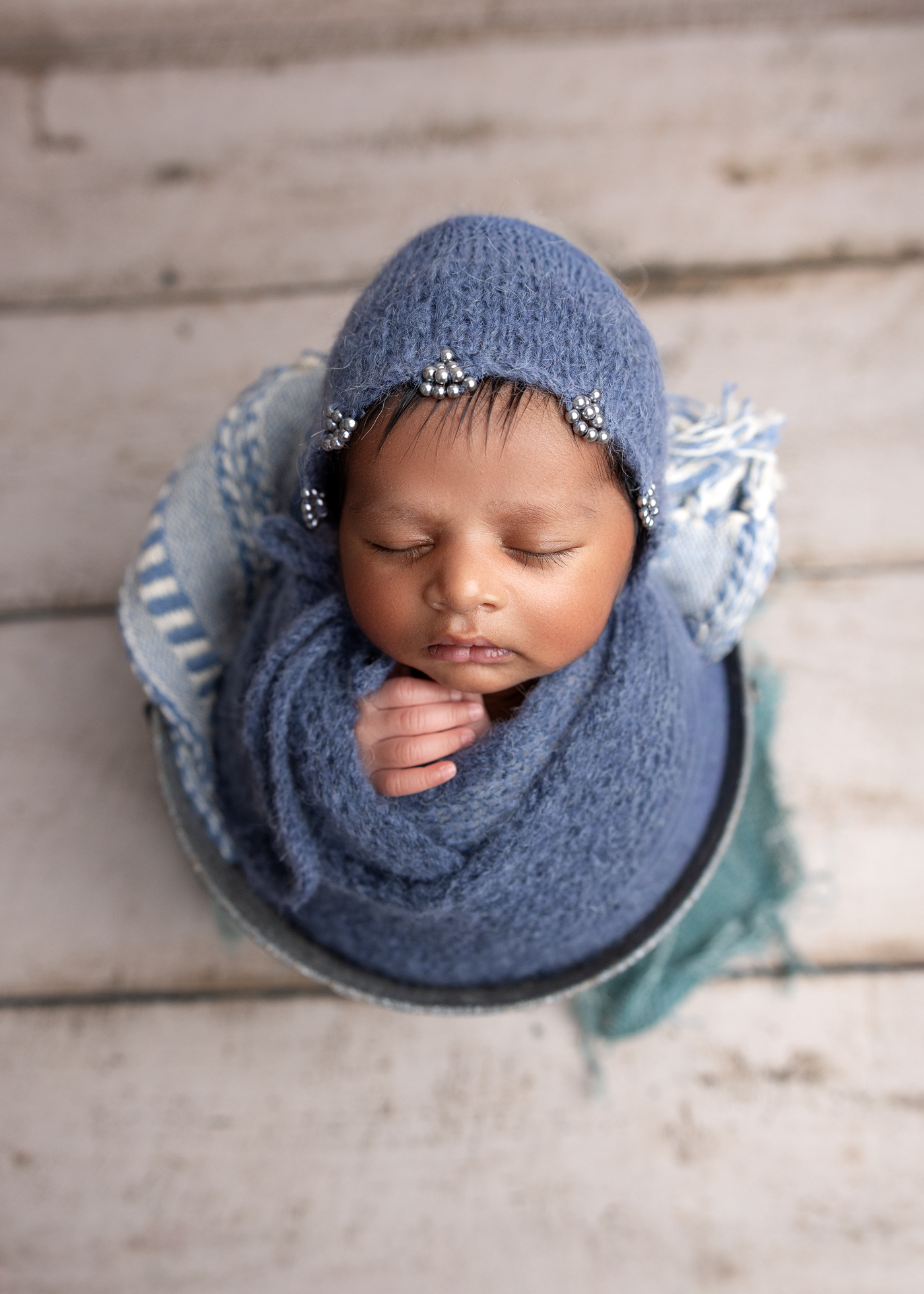 Sleeping newborn wrapped in soft blue knit blanket and bonnet rests peacefully in oval basket on wooden floor.