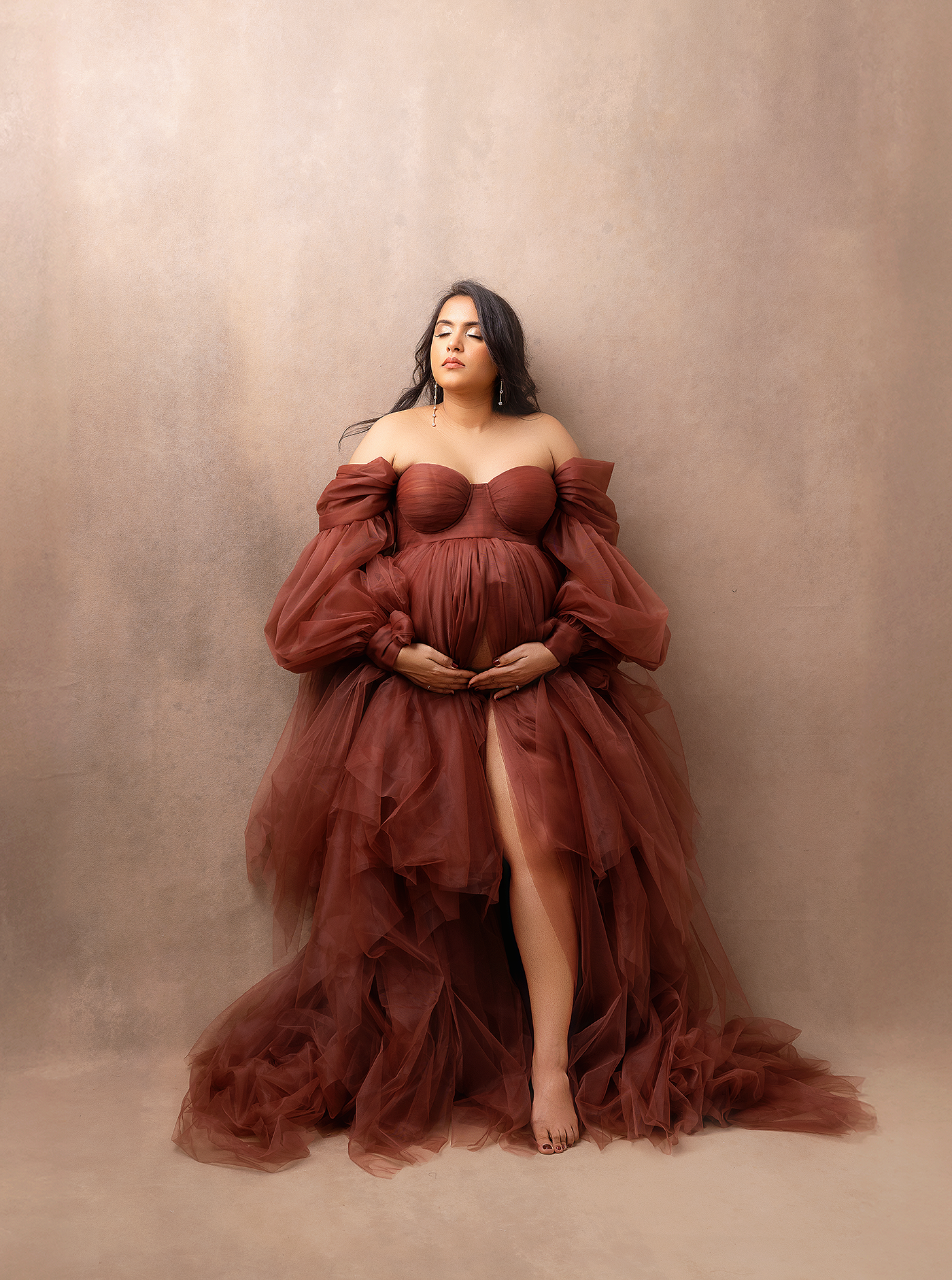 Pregnant woman in elegant rust tulle off-shoulder maternity gown poses gracefully in professional studio photo.