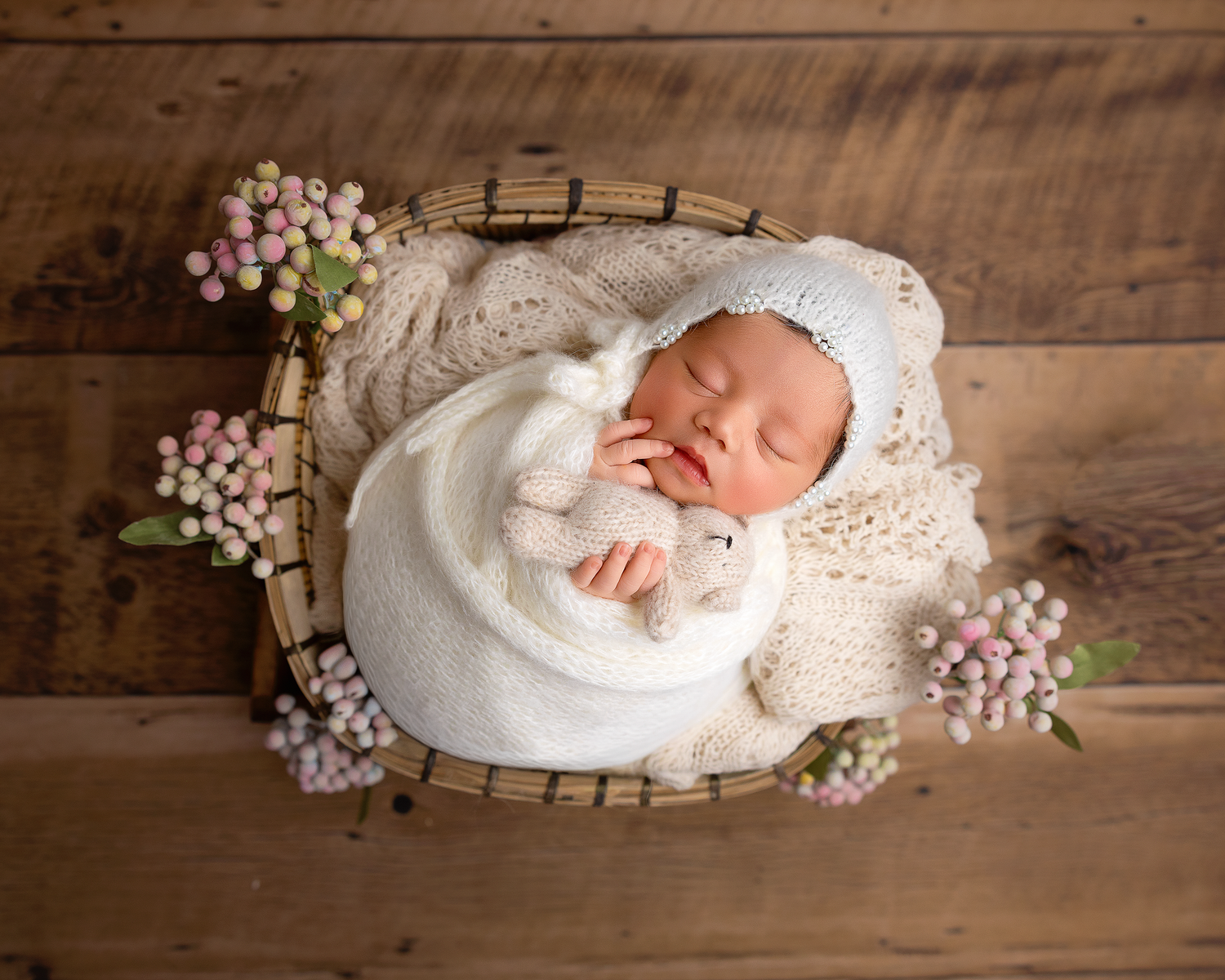 A sleeping newborn baby wrapped in white lace fabric lies on rustic wooden boards with delicate pink flowers.