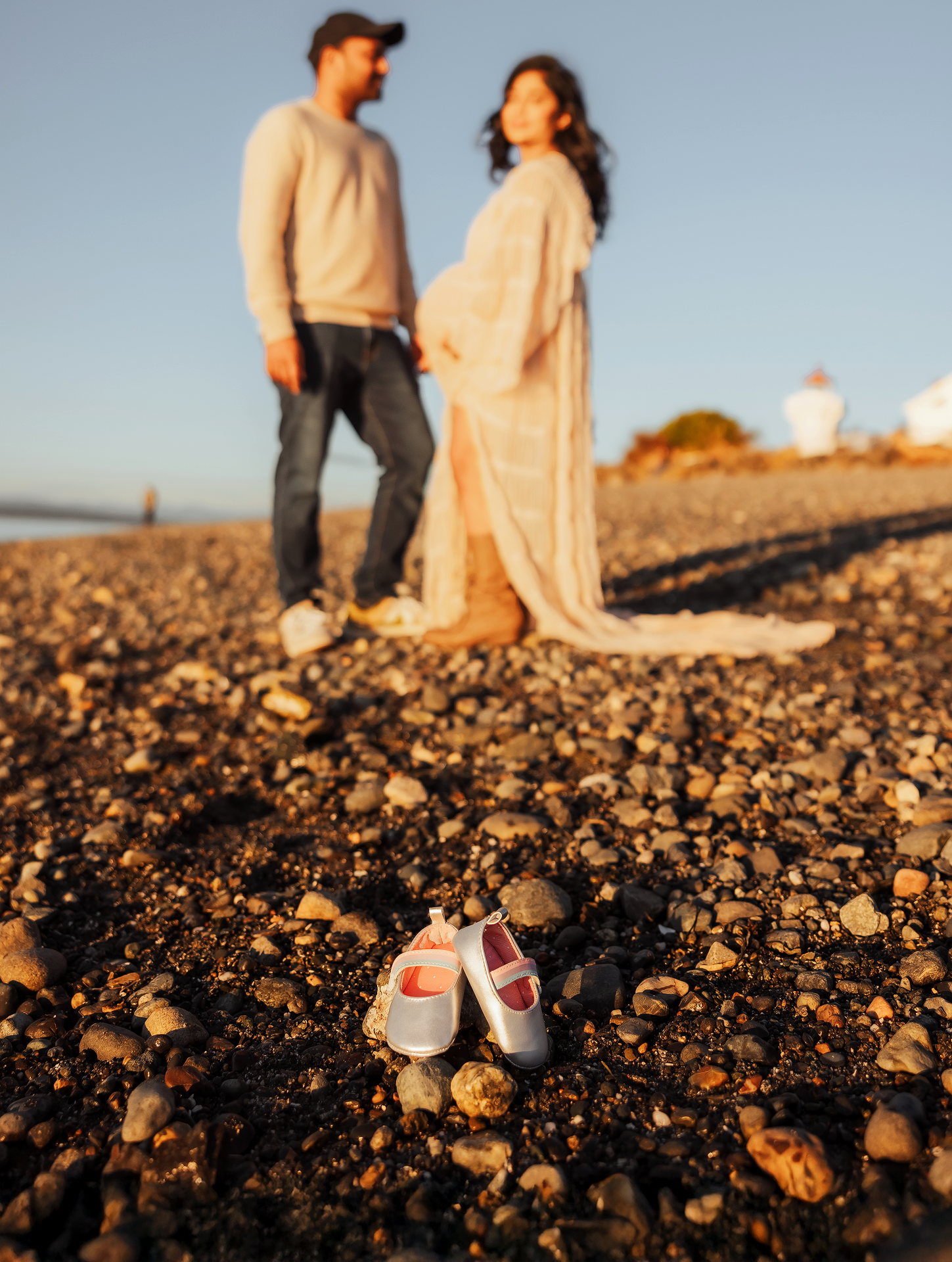 Couple standing on a rocky beach during golden hour with tiny baby shoes in the foreground at a maternity photoshoot.