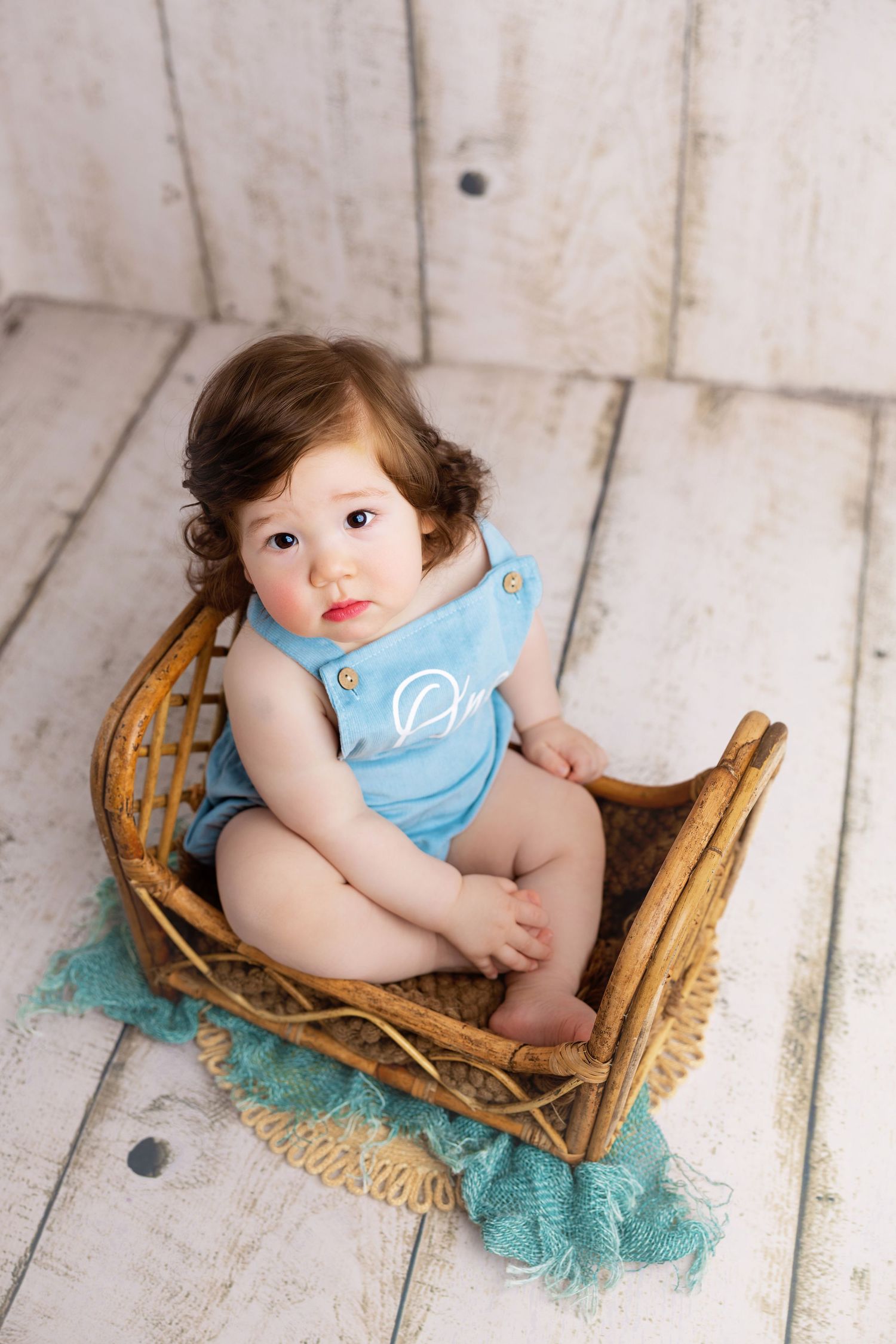 Adorable baby in blue monogrammed romper sitting in a wicker basket on white wood floor with teal blanket.