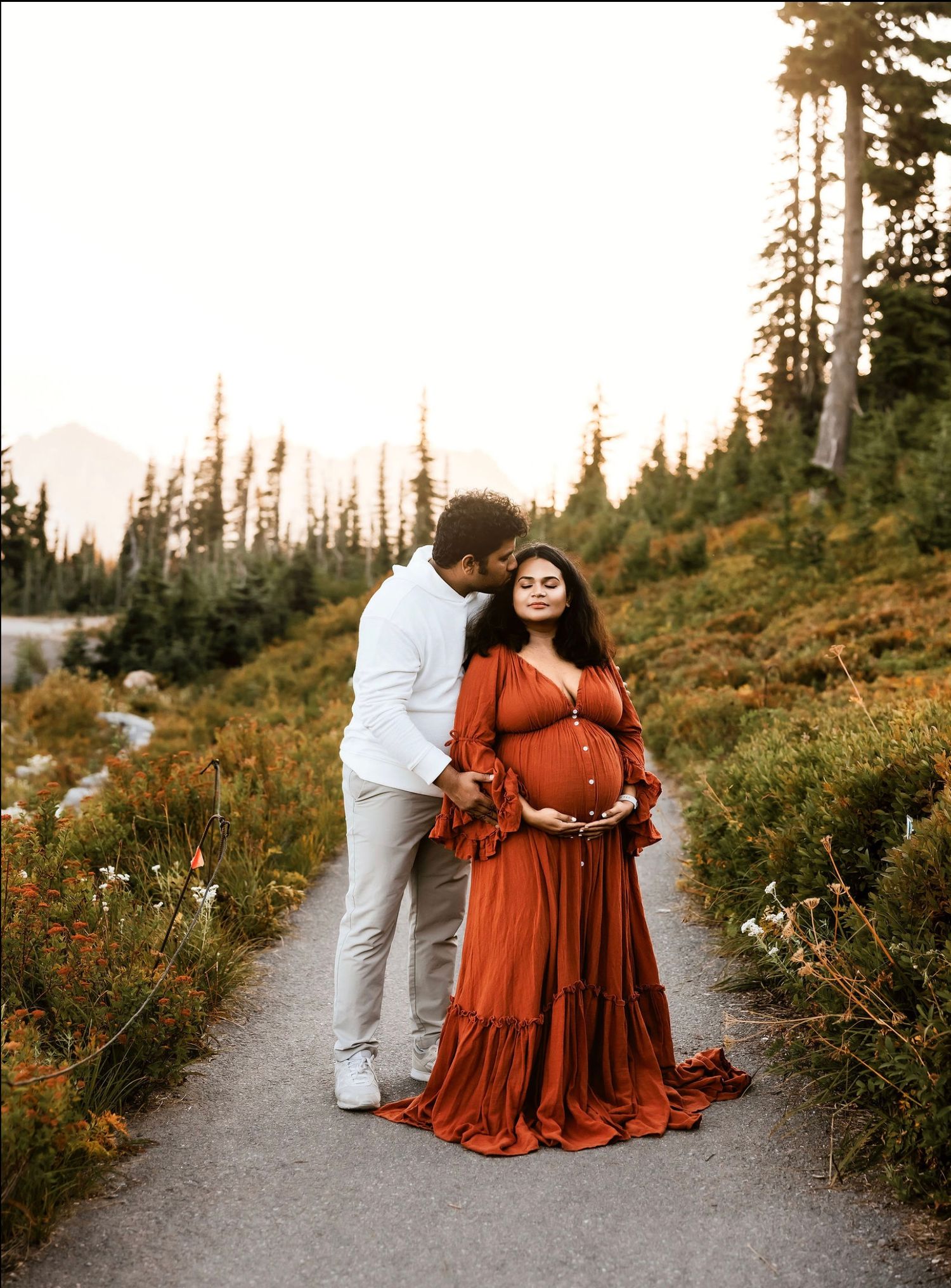 A couple poses together on a mountain trail at sunset surrounded by evergreen trees and wildflowers.