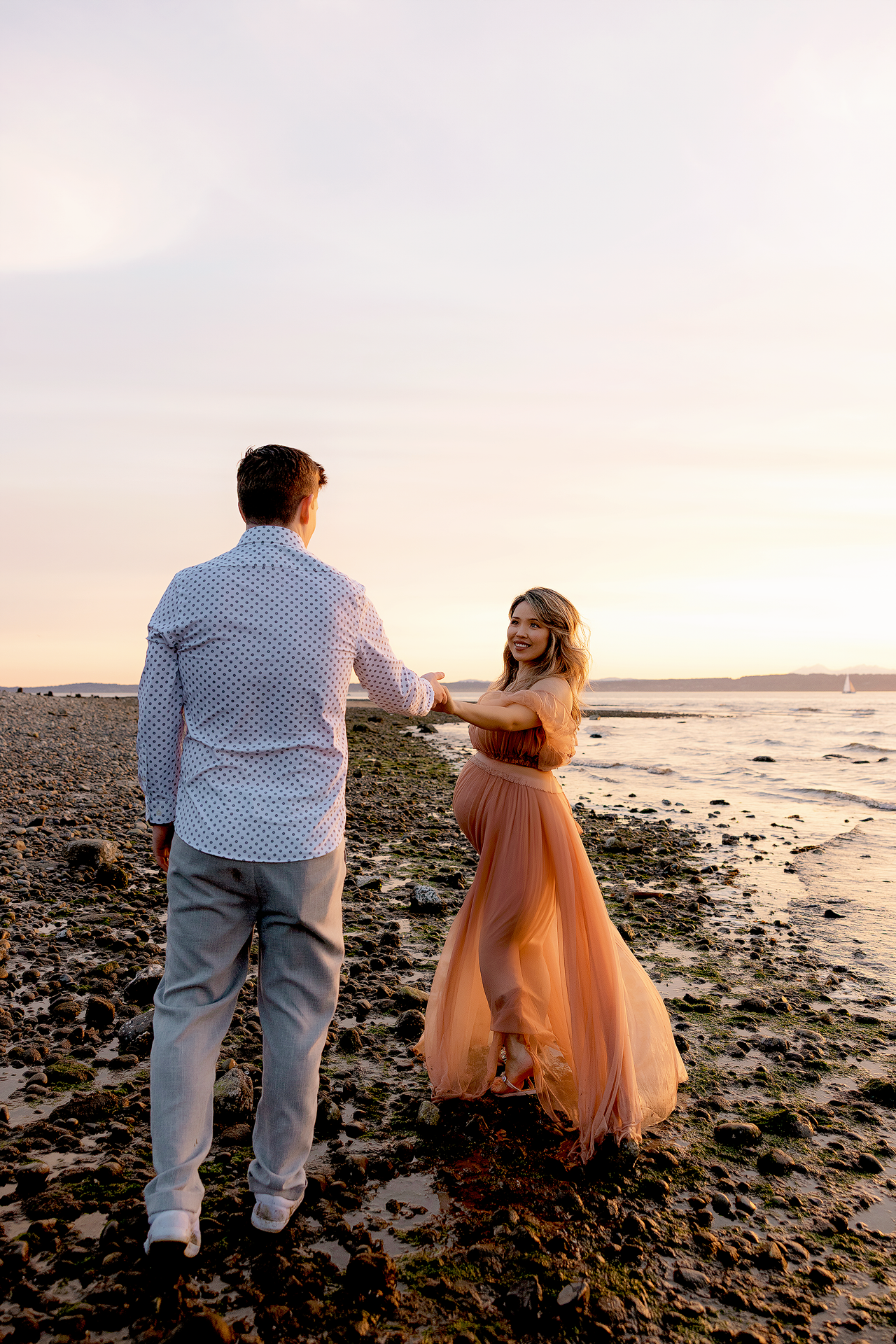 Couple holding hands on rocky beach at sunset, woman in flowing pink gown smiling at partner in white shirt.