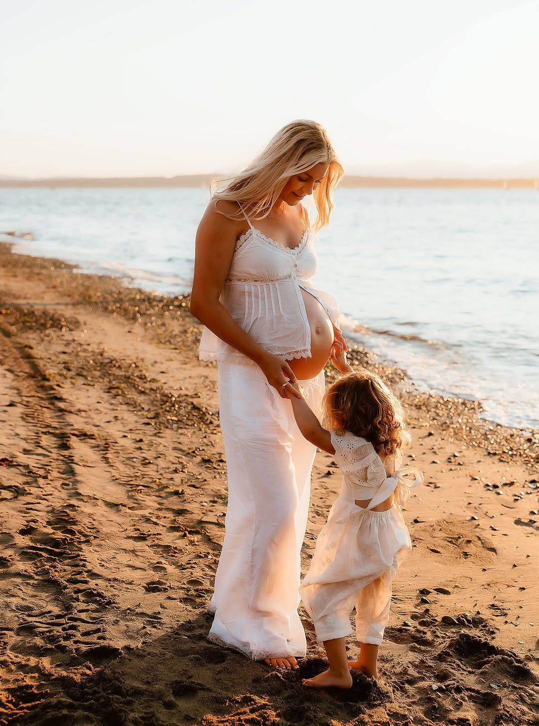 Pregnant woman in white dress holding hands with toddler on sandy beach at golden hour sunset.