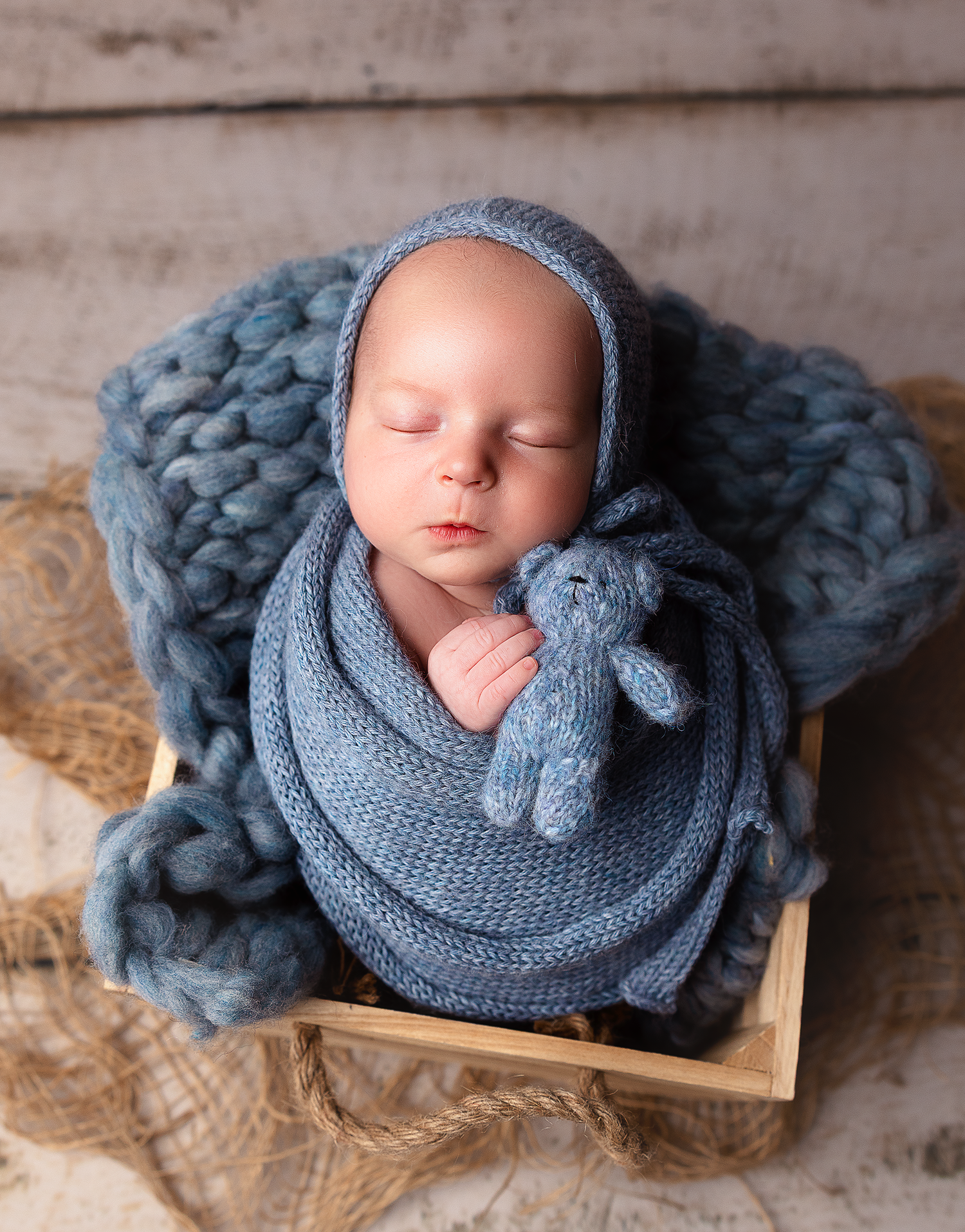 A sleeping newborn swaddled in soft blue knit wraps and blankets rests in a wooden box with a small stuffed toy.