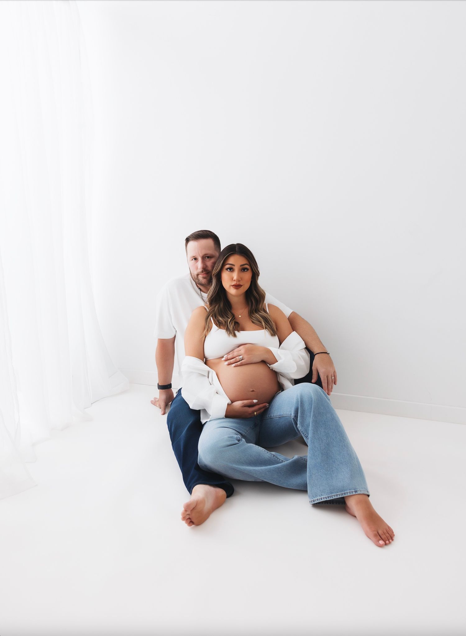 Couple maternity photo shoot, pregnant woman in white top and jeans sitting with partner against white studio backdrop.