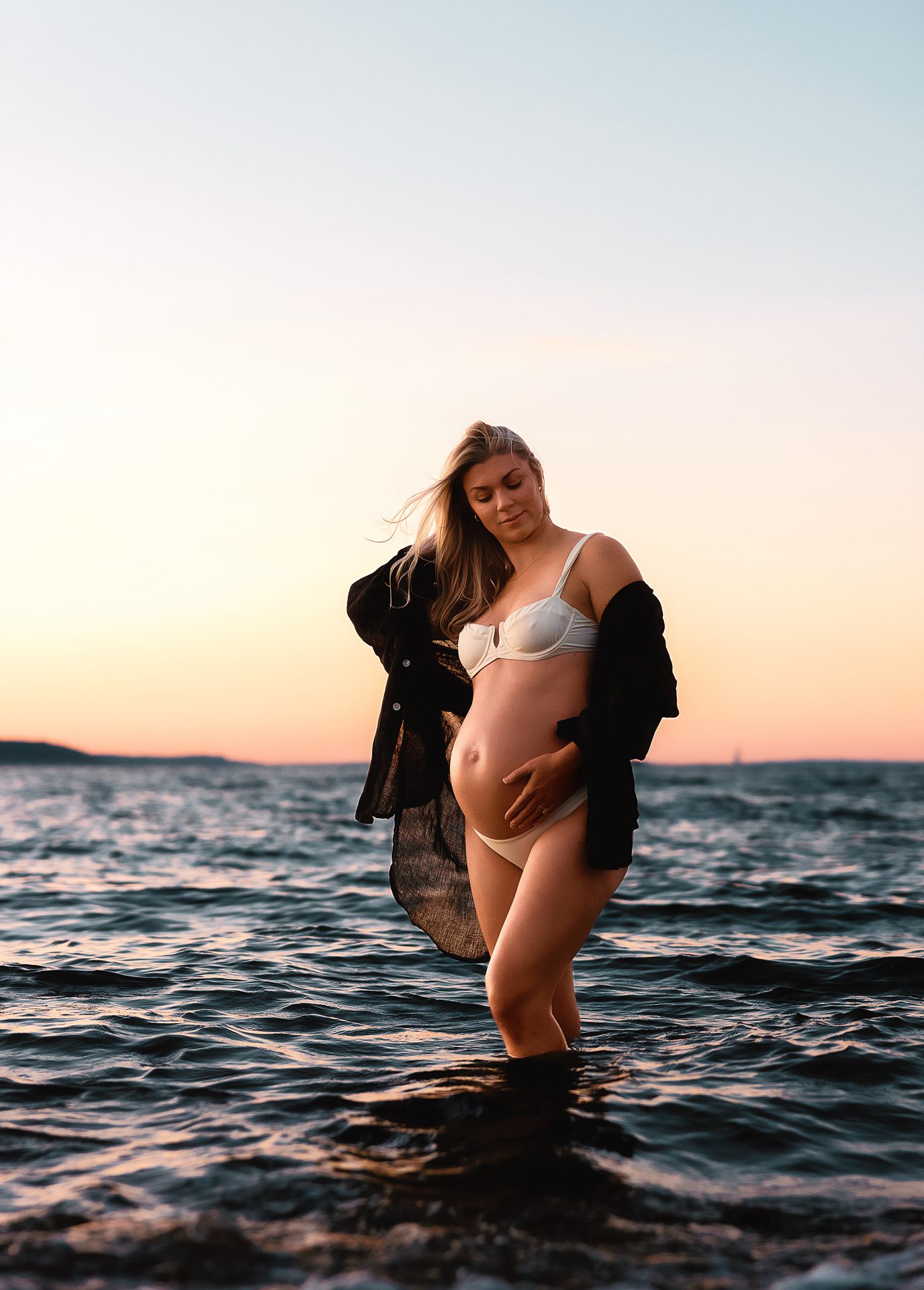 Pregnant woman in white bikini and black sheer jacket standing in ocean water at sunset.