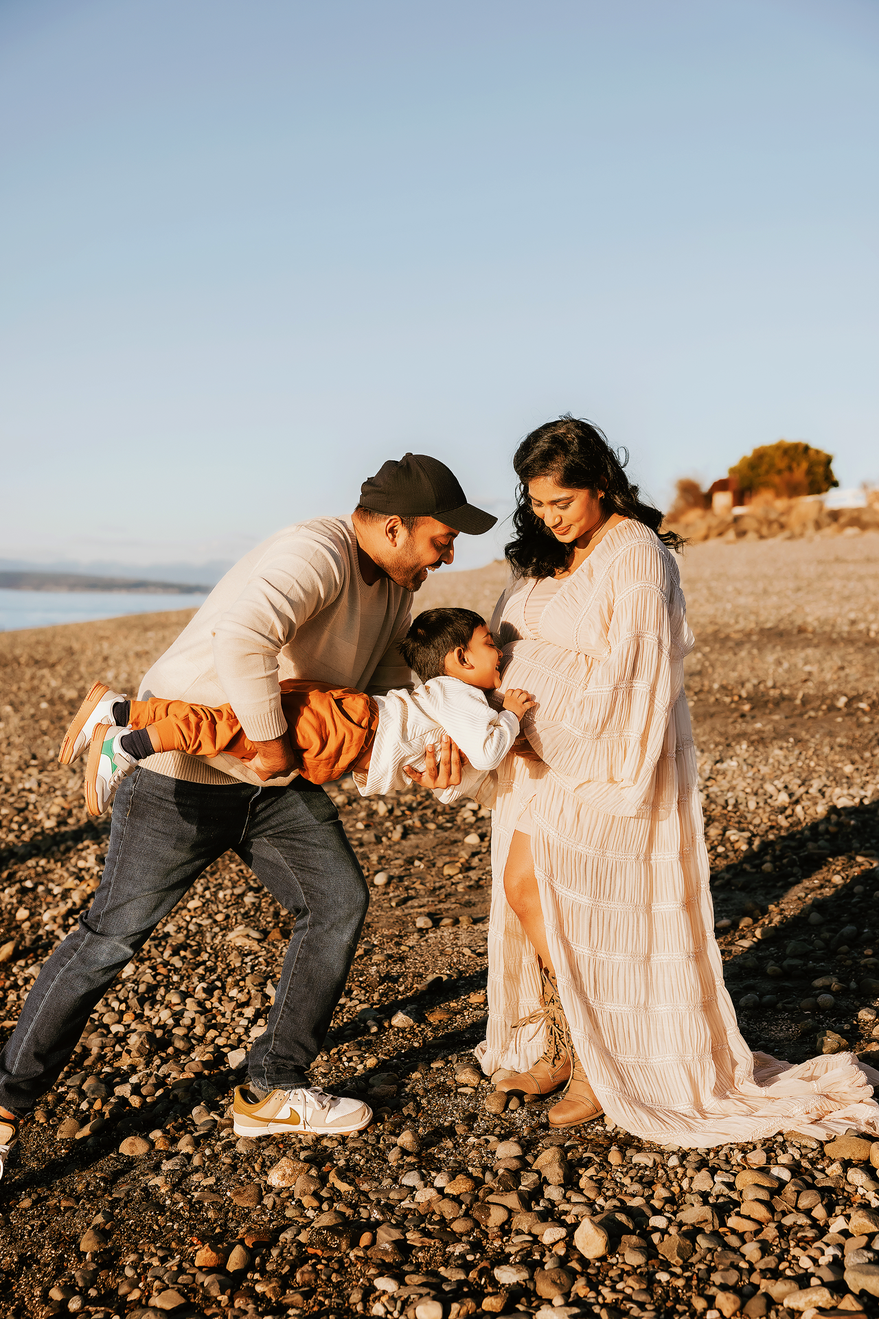 A family of three enjoys a golden hour beach moment, with dad holding a baby toward pregnant mom on a rocky shore.