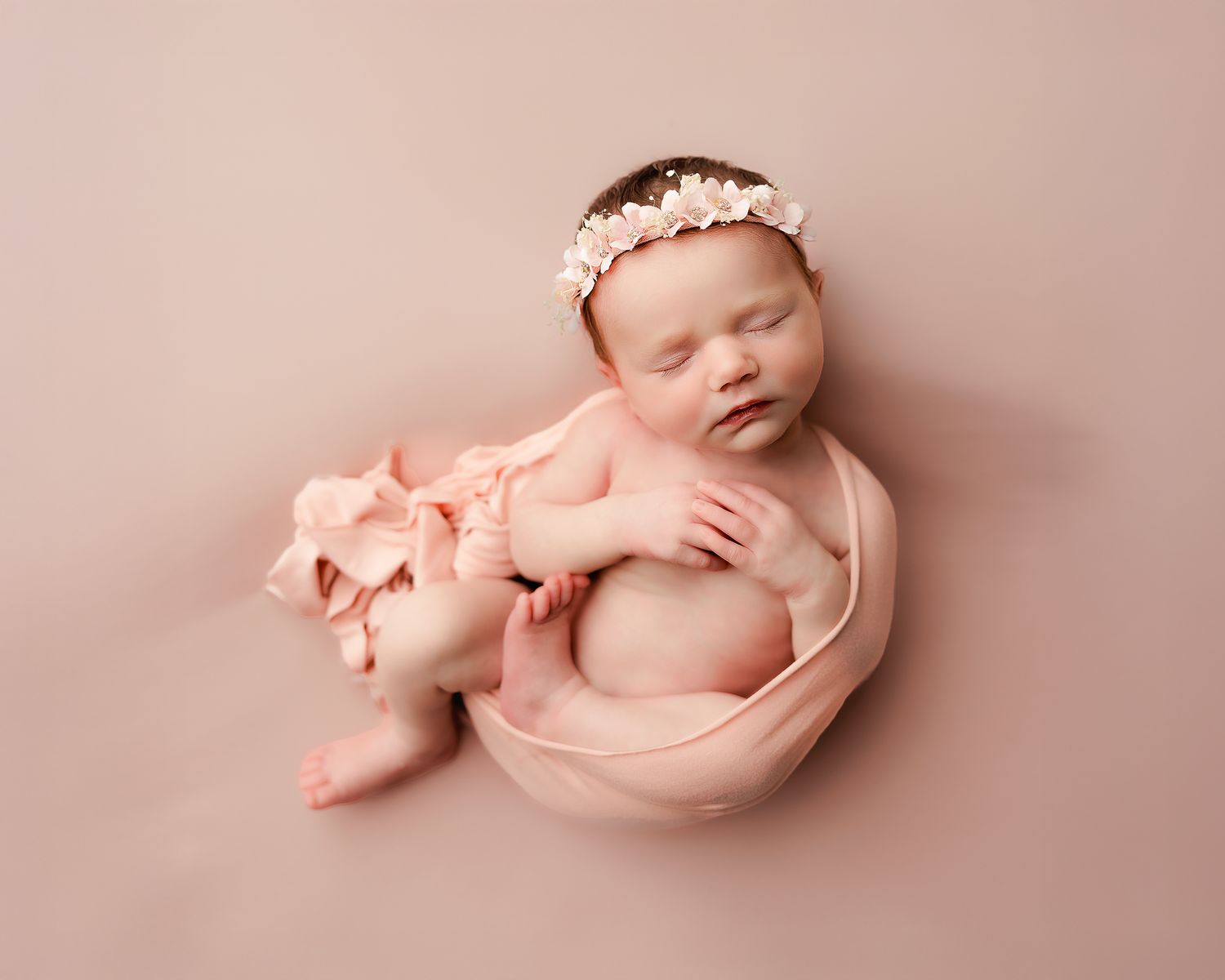Sleeping newborn wrapped in soft pink fabric with matching headband rests peacefully on pink background.
