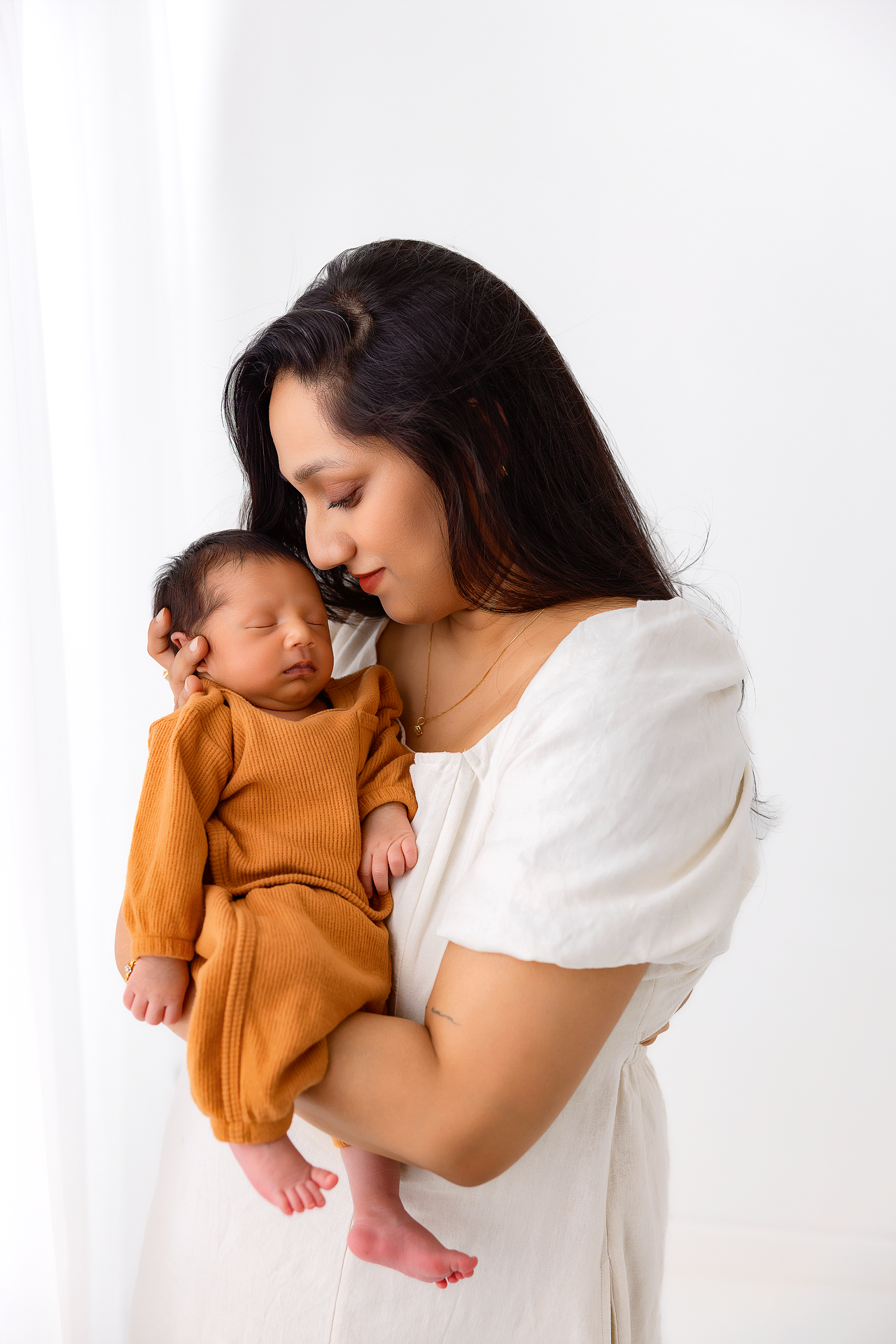 A tender moment between a parent and sleeping newborn baby in a brown onesie against a white background.
