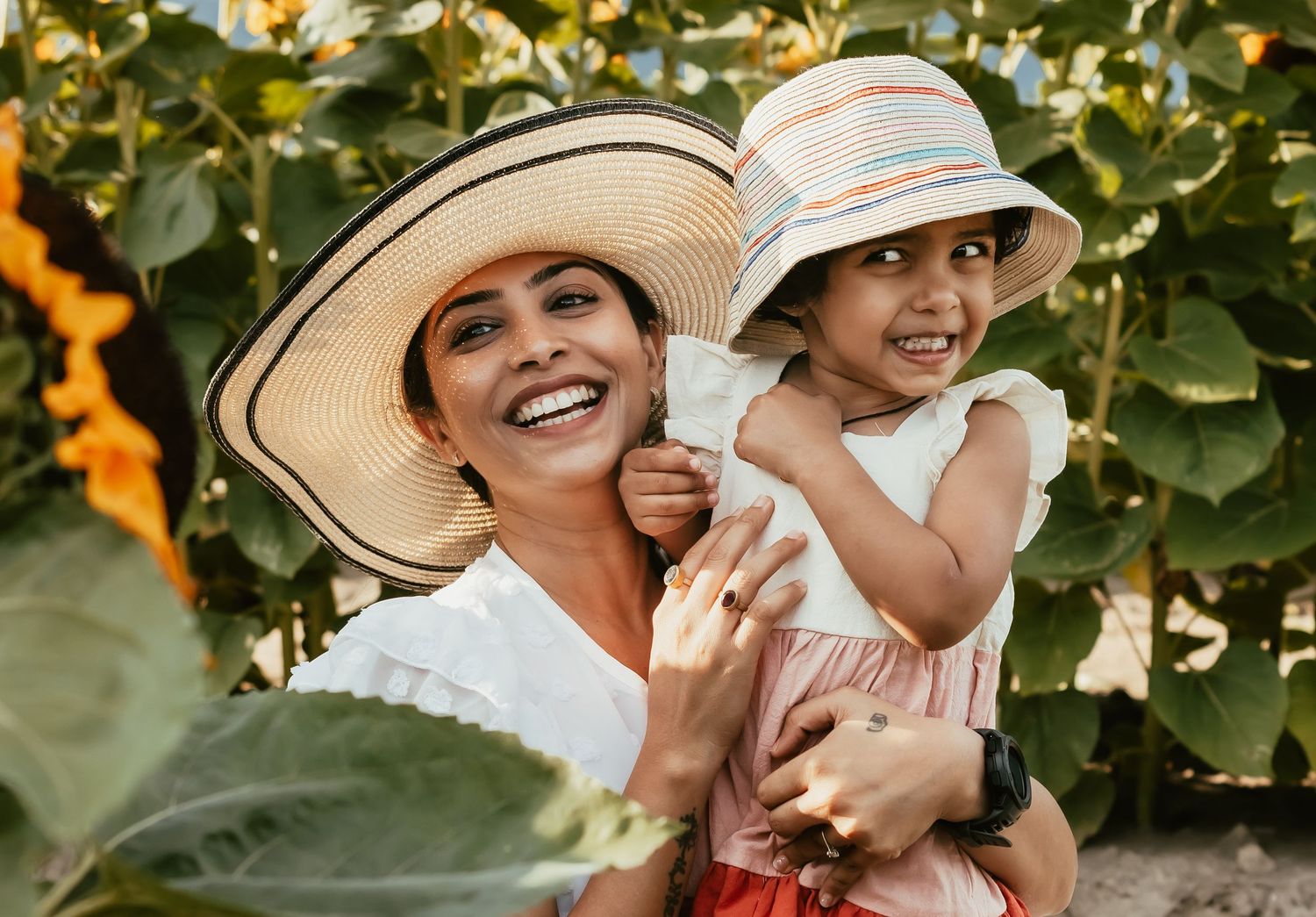 Two people wearing sun hats embrace and laugh together in a sunny vineyard setting.