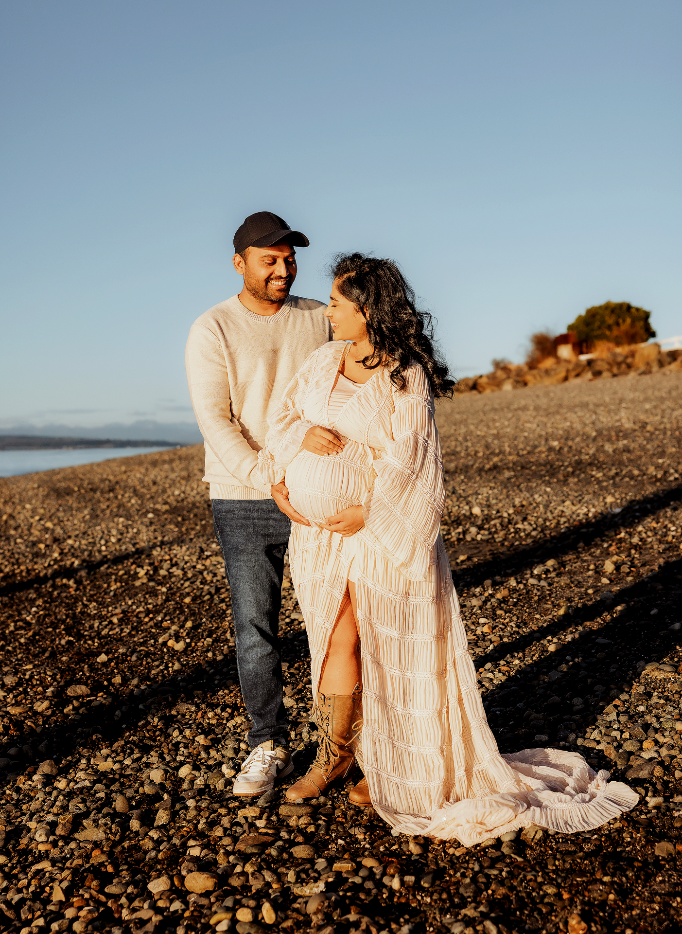 Couple poses on rocky beach at golden hour, pregnant woman in flowing white dress held by partner in beige sweater.