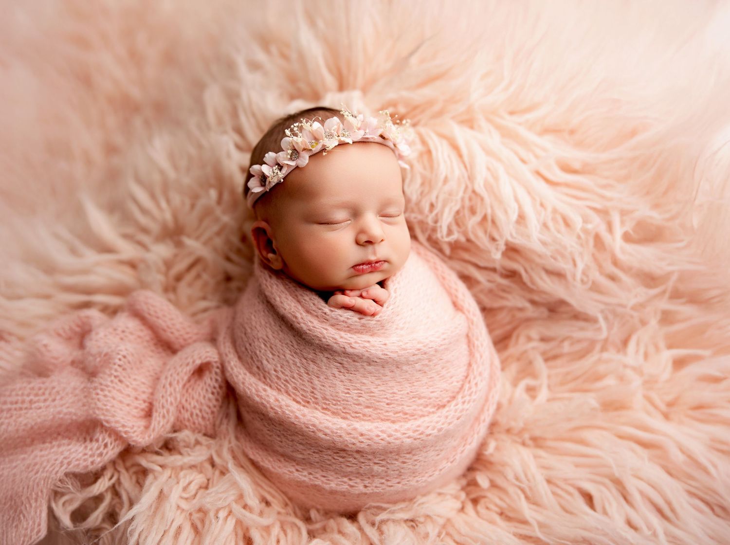 Sleeping newborn baby girl wrapped in pink swaddle with floral headband on fluffy pink fur backdrop.