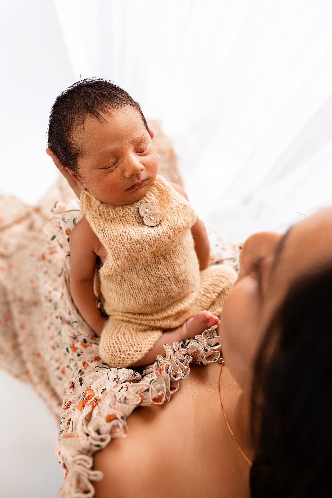 A tender moment between parent and sleeping newborn baby in warm peach tones against white bedding.