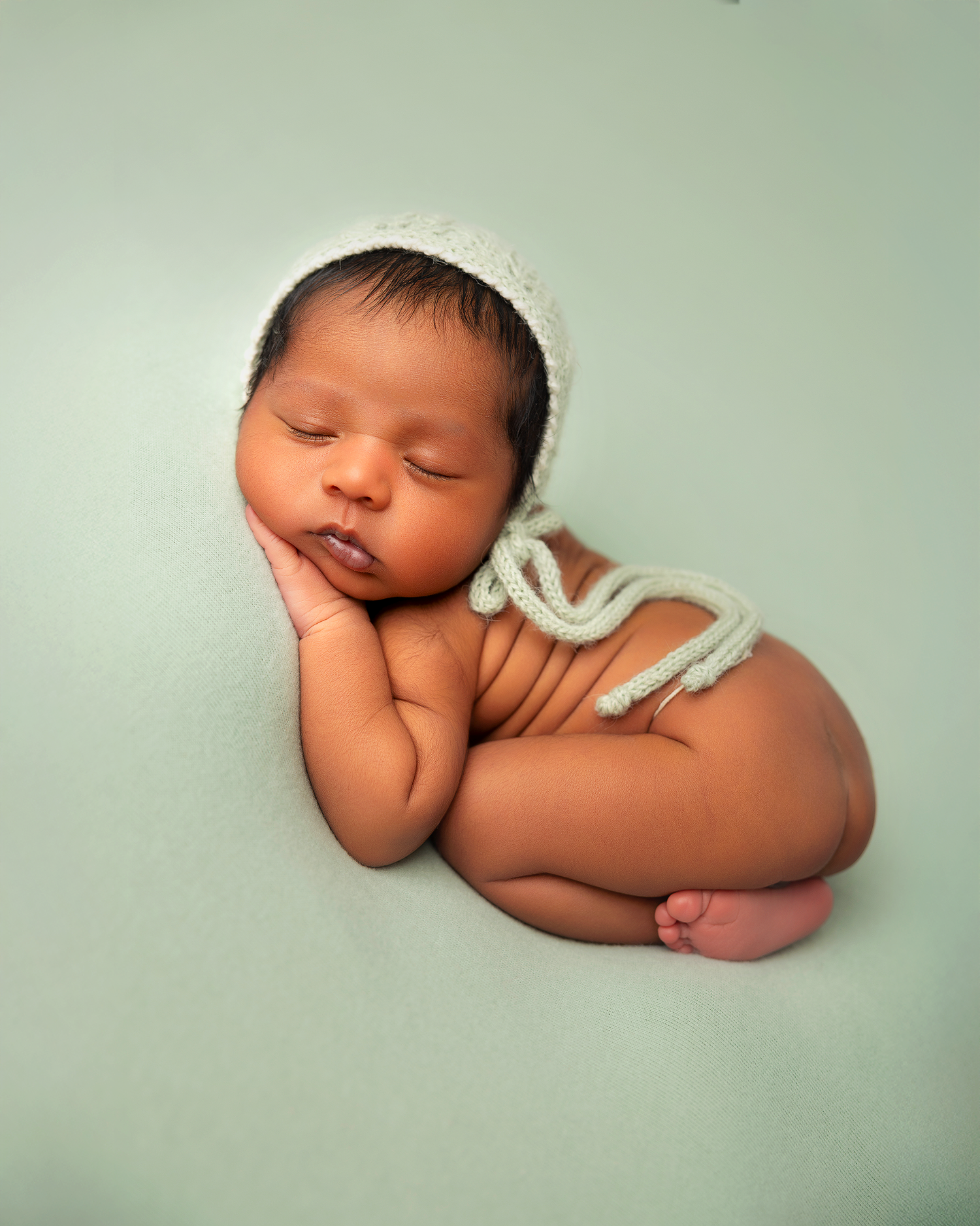 Sleeping newborn baby wearing a white knit bonnet, curled up on a soft mint green background.