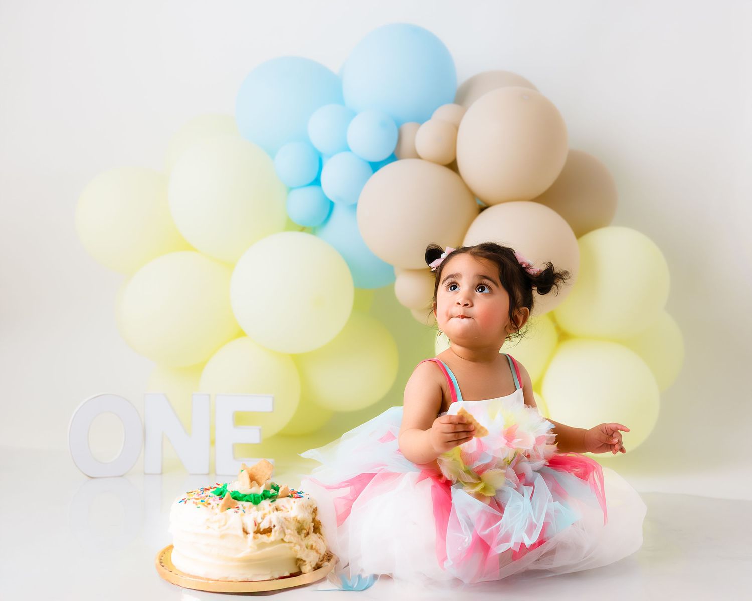 Pastel balloon arch decorates cake smash photo setup with pink tutu and white backdrop.