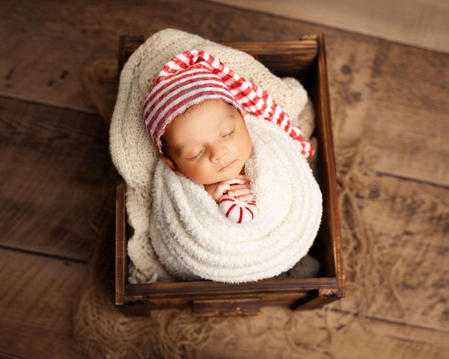 Sleeping newborn baby wearing red and white striped hat nestled in wooden box with white blanket.