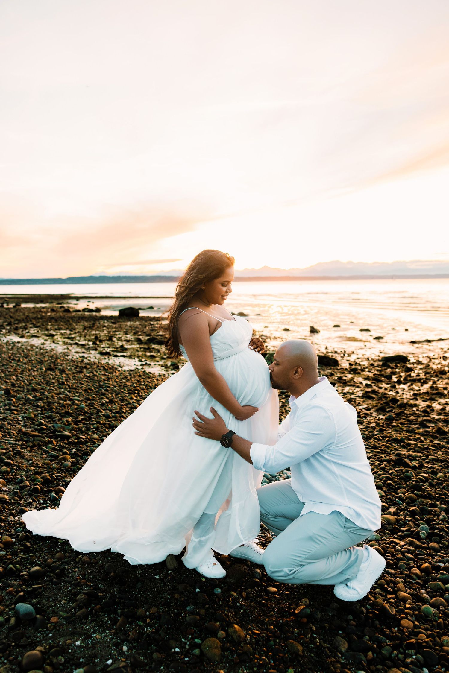 A romantic couple in white attire sits together on a rocky beach at sunset sharing an intimate moment.