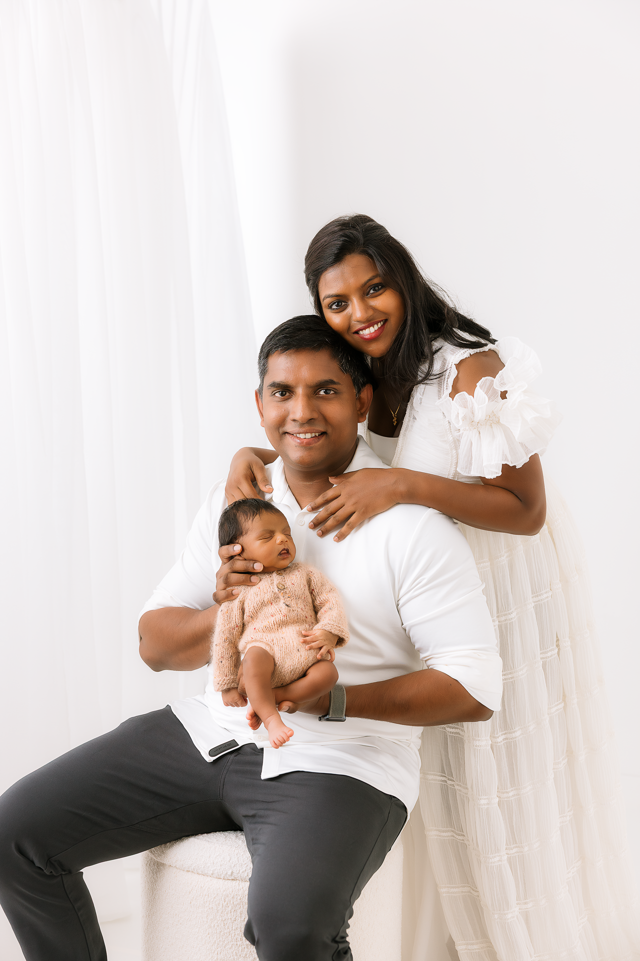 Happy family in white shirts and black pants pose together with their baby against a white background.