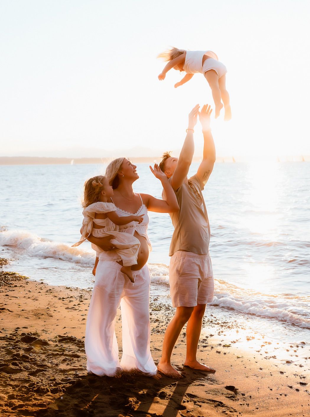 Pregnant woman and man tossing child in air on sunny beach at golden hour, joyful family photo.