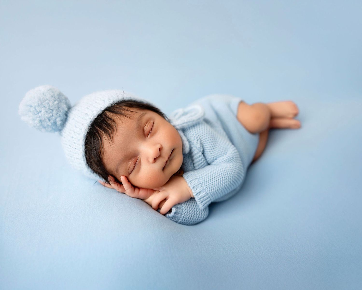 Sleeping newborn baby in blue knit outfit and pom-pom hat on blue background.