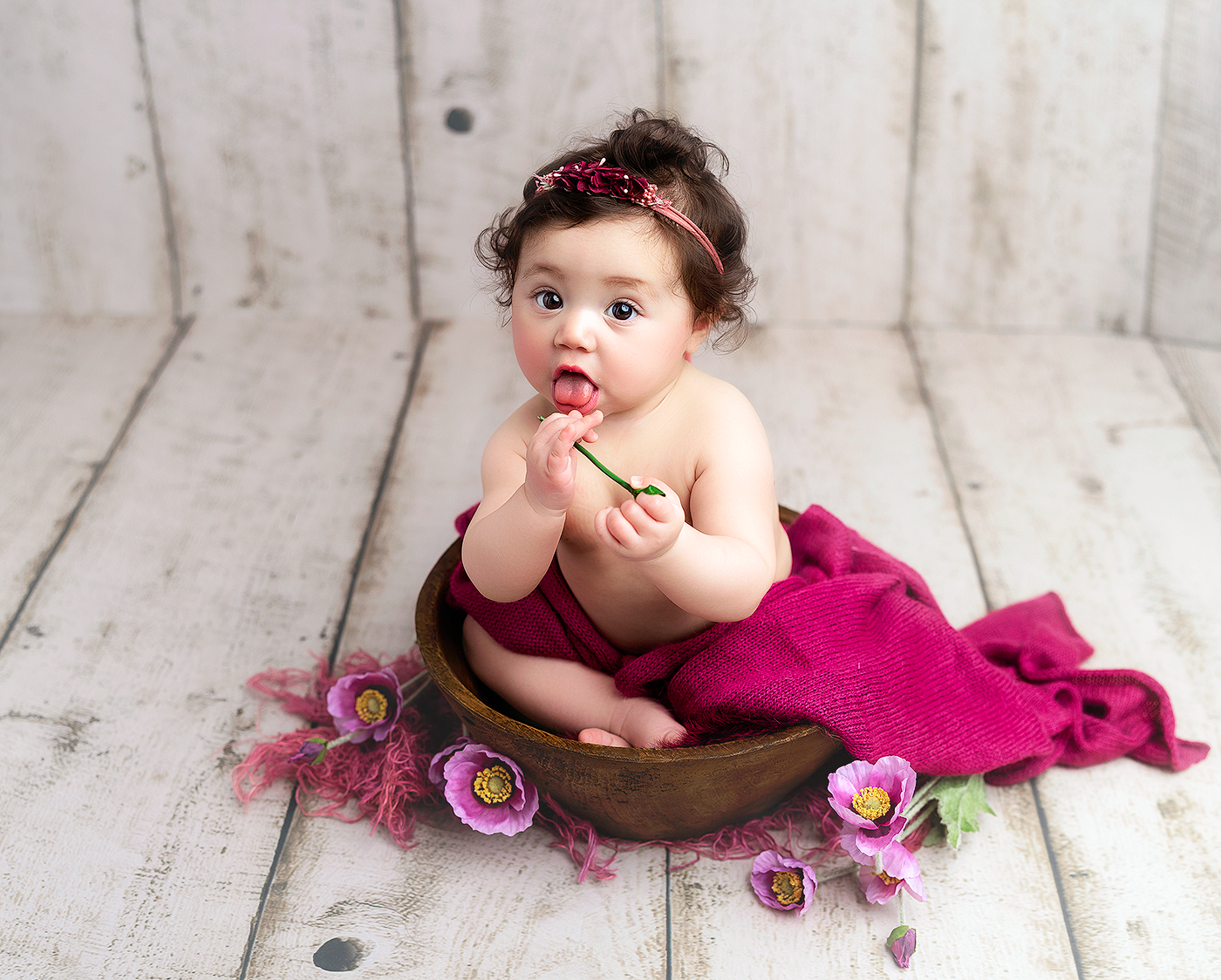 Adorable baby girl sitting in wooden bowl with pink floral wrap and flower headband on rustic white wood floor.