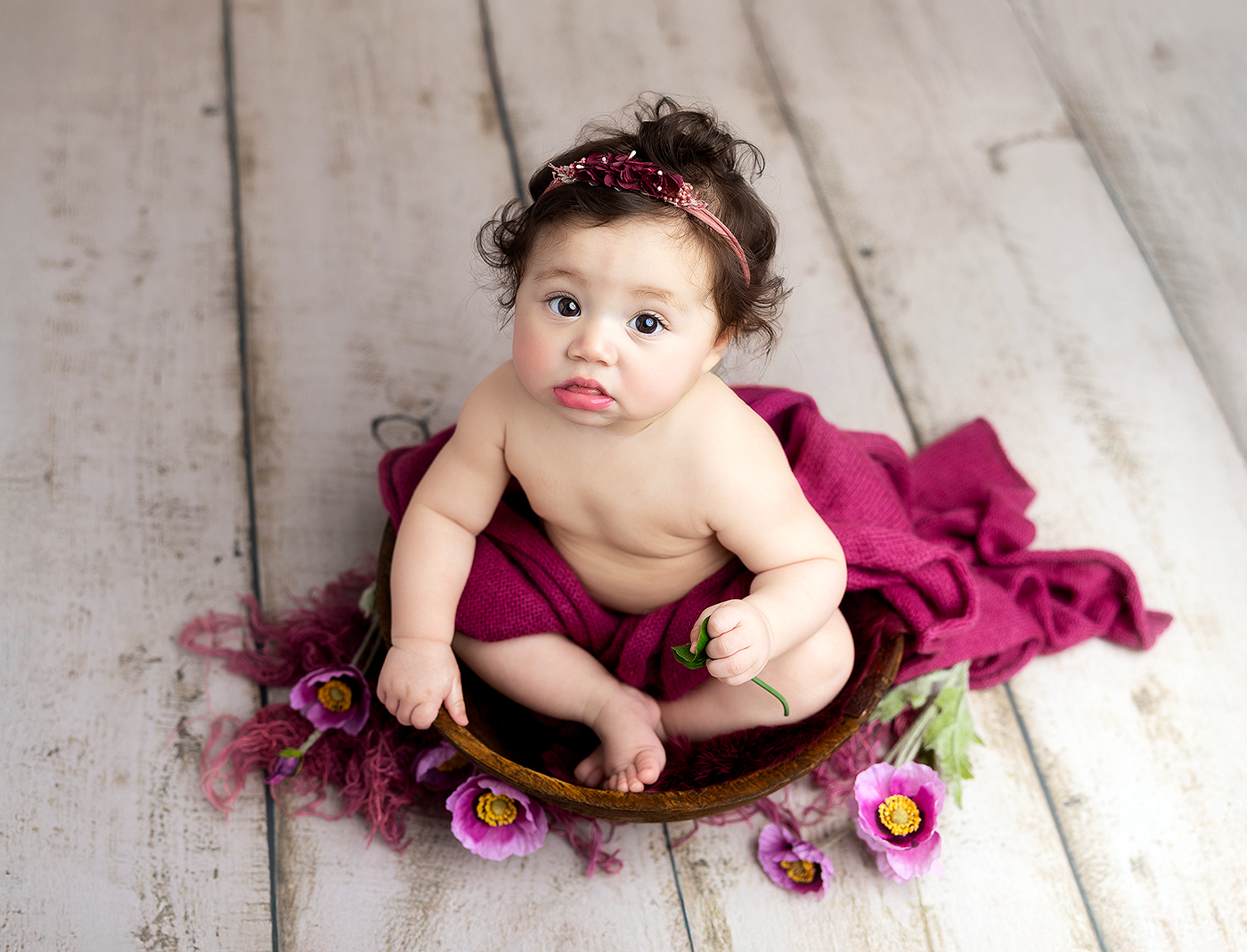 Adorable baby girl in magenta bloomers sits in wooden bowl with purple flowers and matching headband on white wood floor.