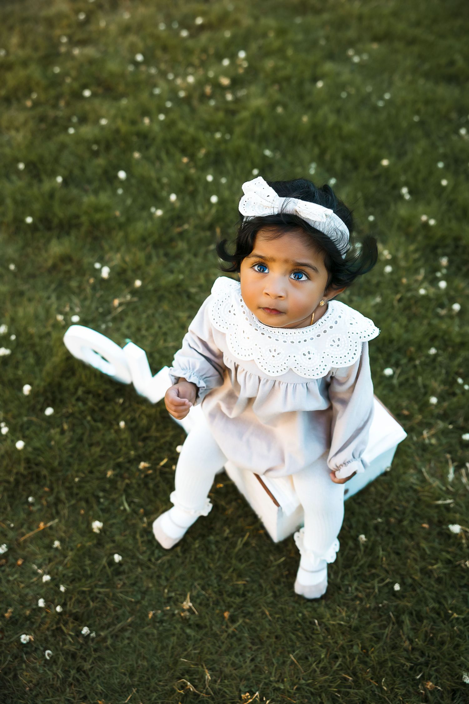 Small child in white lace dress plays on grass with small white flowers.