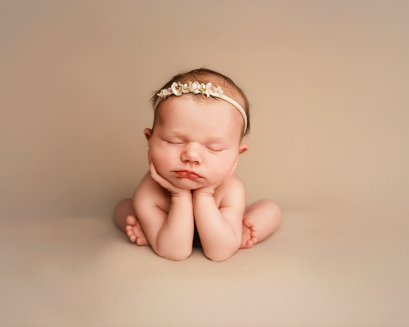 Sleeping newborn baby girl posed with hands under chin wearing a delicate floral headband on a beige background.