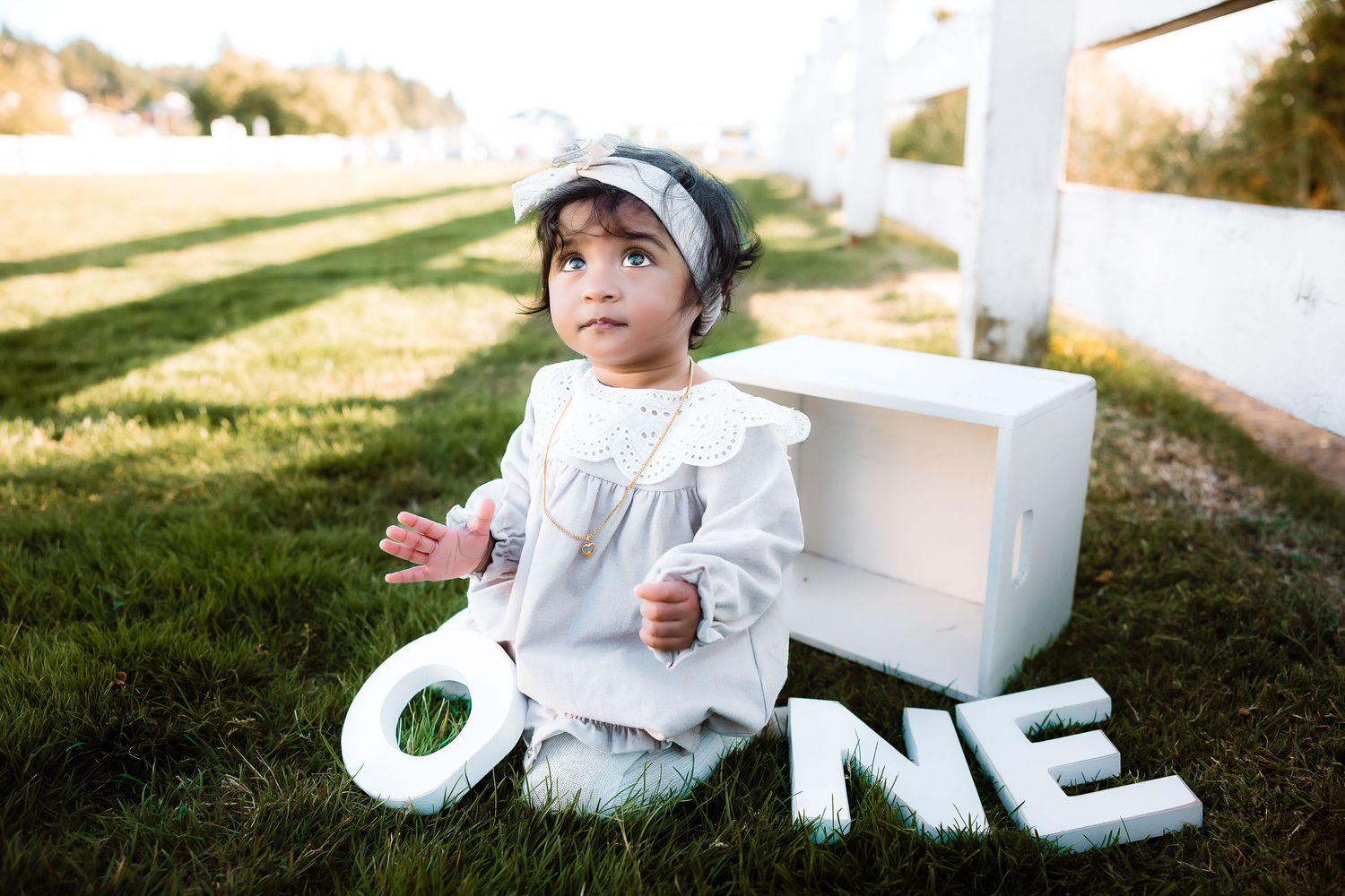 Baby wearing white dress and headband plays on grass with decorative white letters.