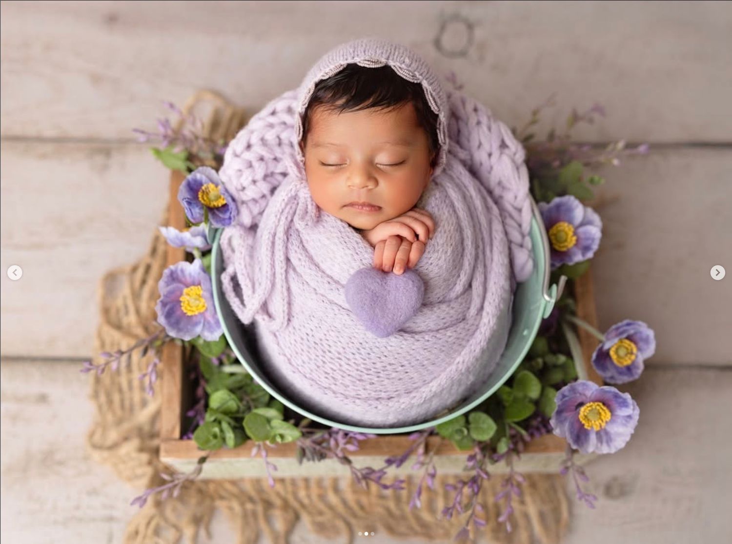 Newborn baby sleeping peacefully in a lavender knit wrap inside a mint bucket with purple flowers.