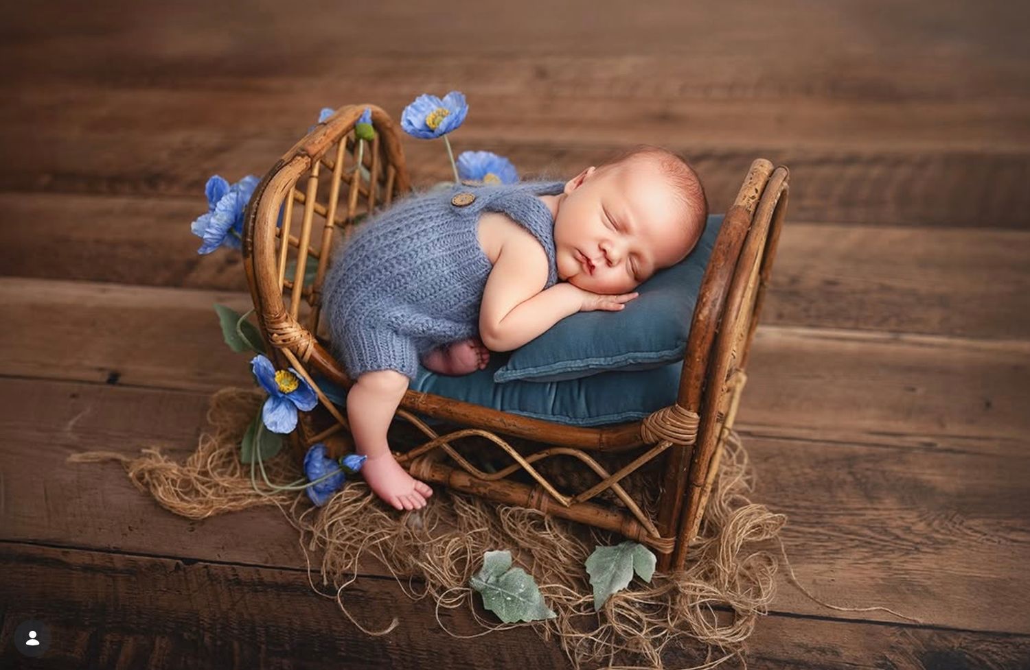 Newborn baby sleeping peacefully in a rustic wicker bed with blue flowers and a teal pillow.