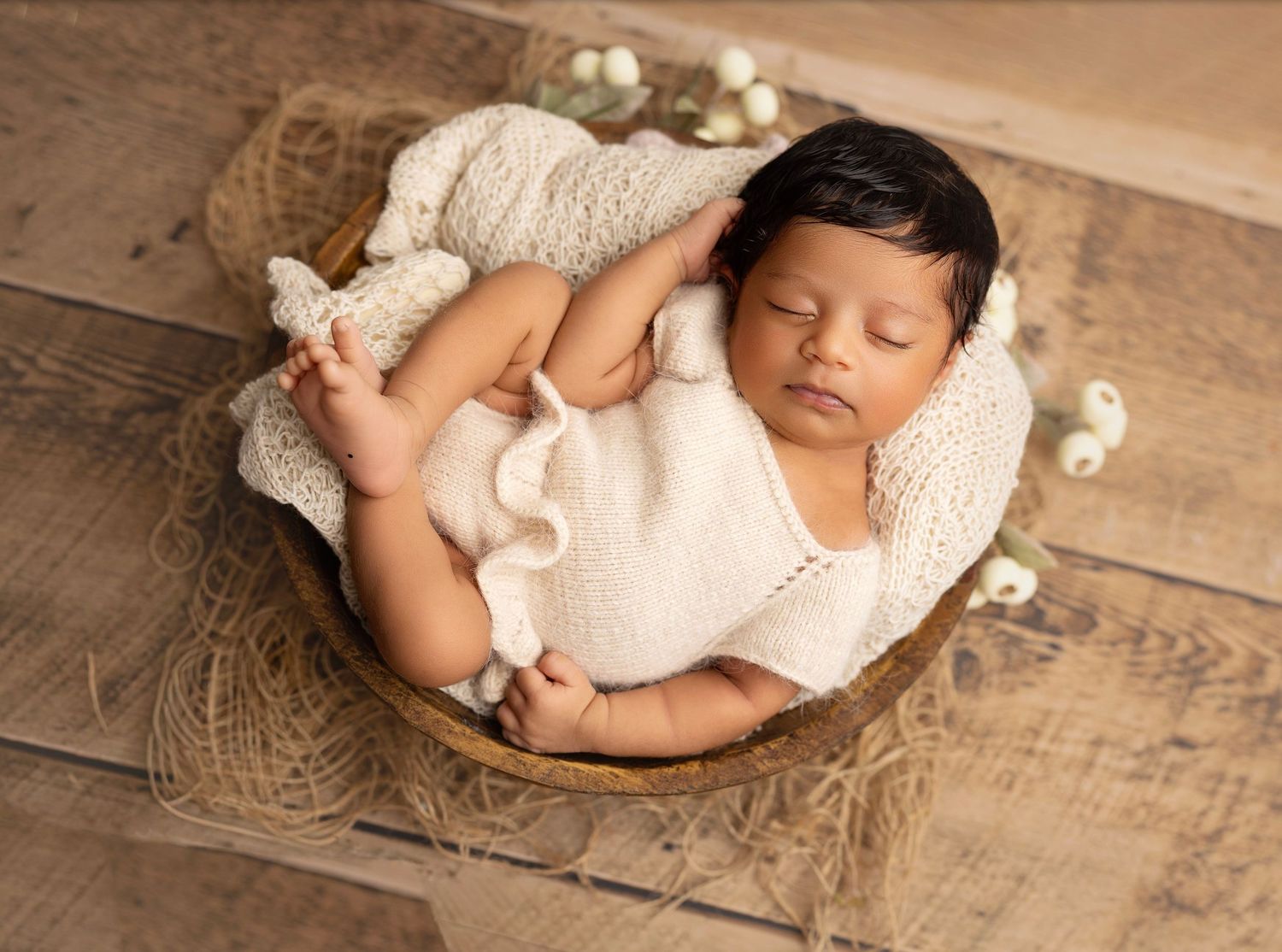 Sleeping newborn baby in cream knit outfit nestled in a rustic wooden bowl on hardwood floor.