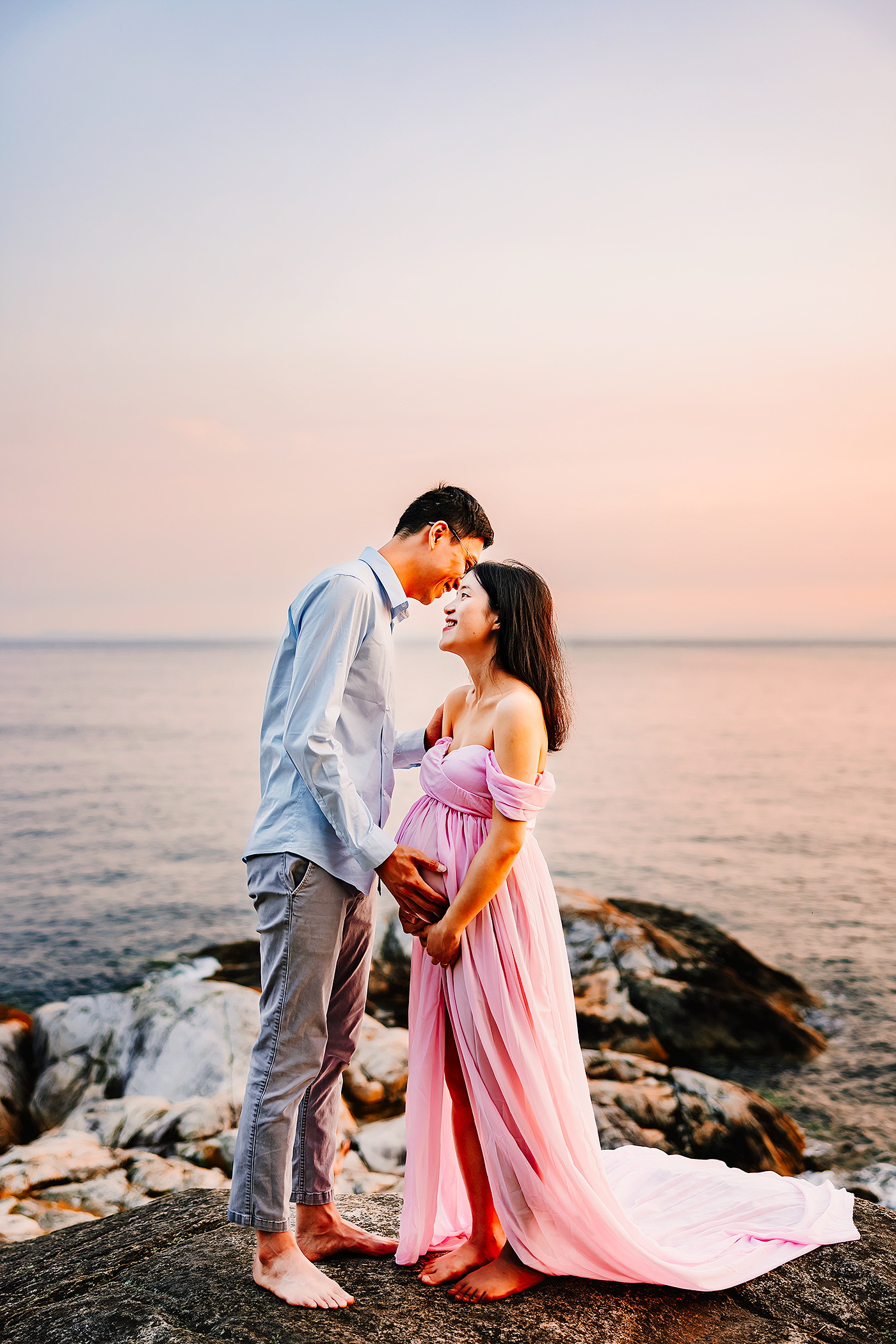 A romantic couple embraces on coastal rocks at sunset, with the woman wearing a flowing pink dress against an ocean backdrop.
