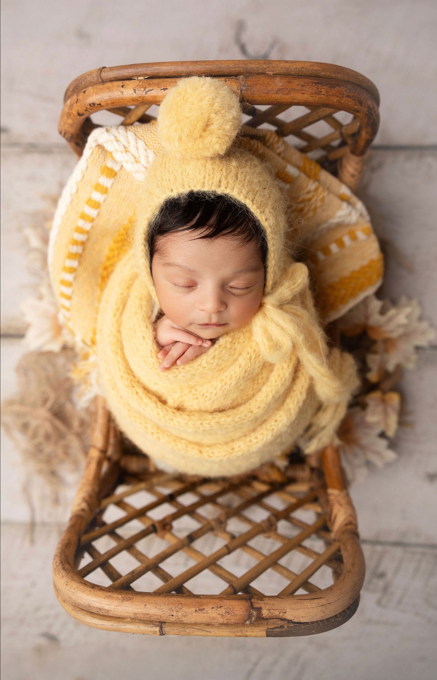 A creative newborn photography setup showing a baby nestled in a wooden basket with a yellow knit blanket.