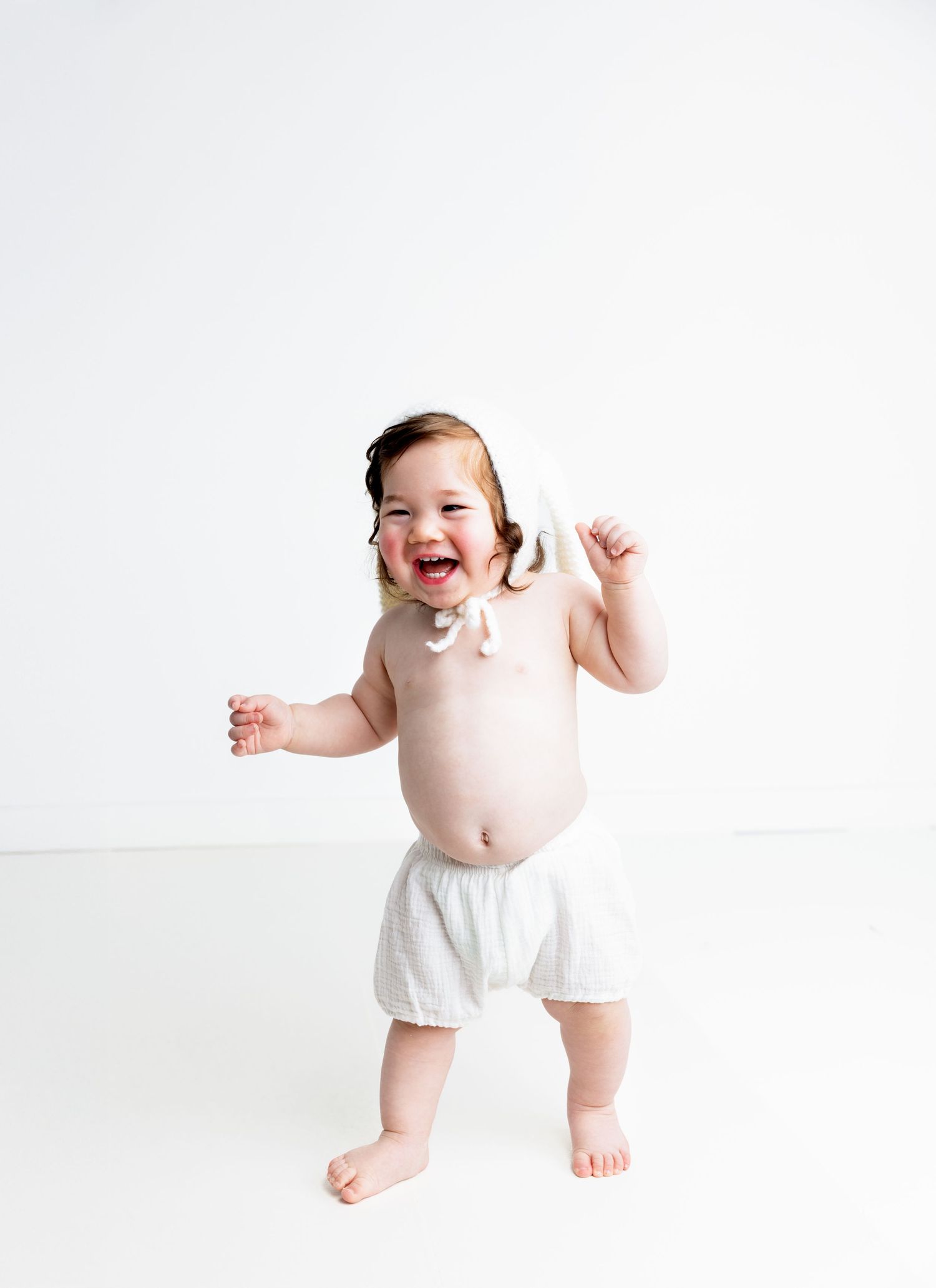 Happy baby in white bloomers and bow laughing while standing on white background.