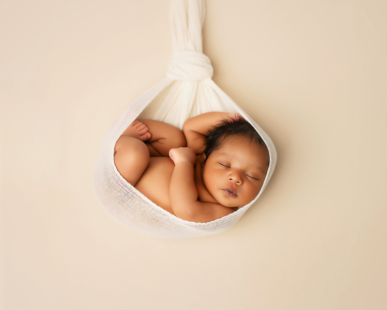 Newborn baby sleeping peacefully in a white fabric sling against a soft beige background.