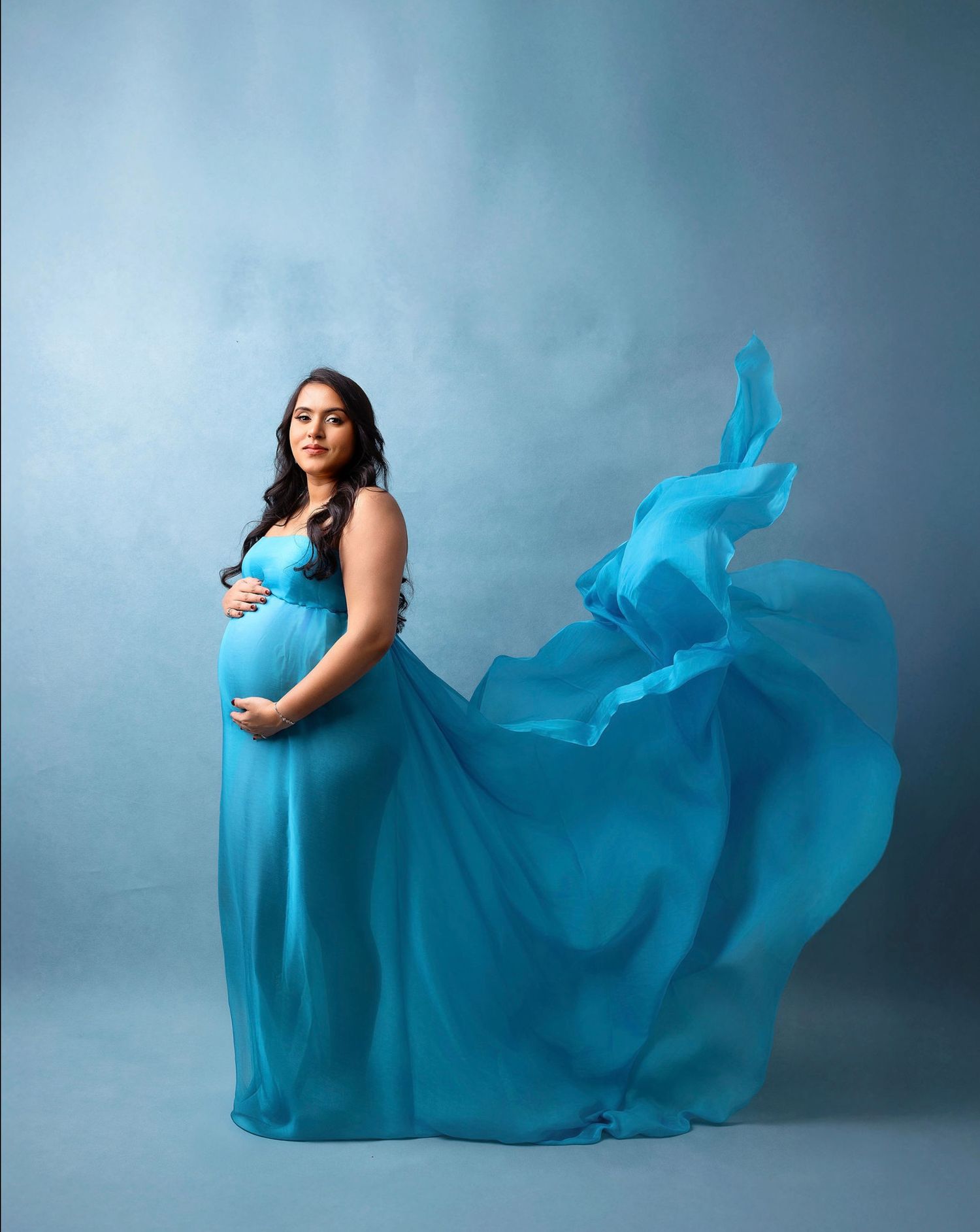 A flowing turquoise maternity dress billows gracefully against a blue studio backdrop in a series of elegant poses.
