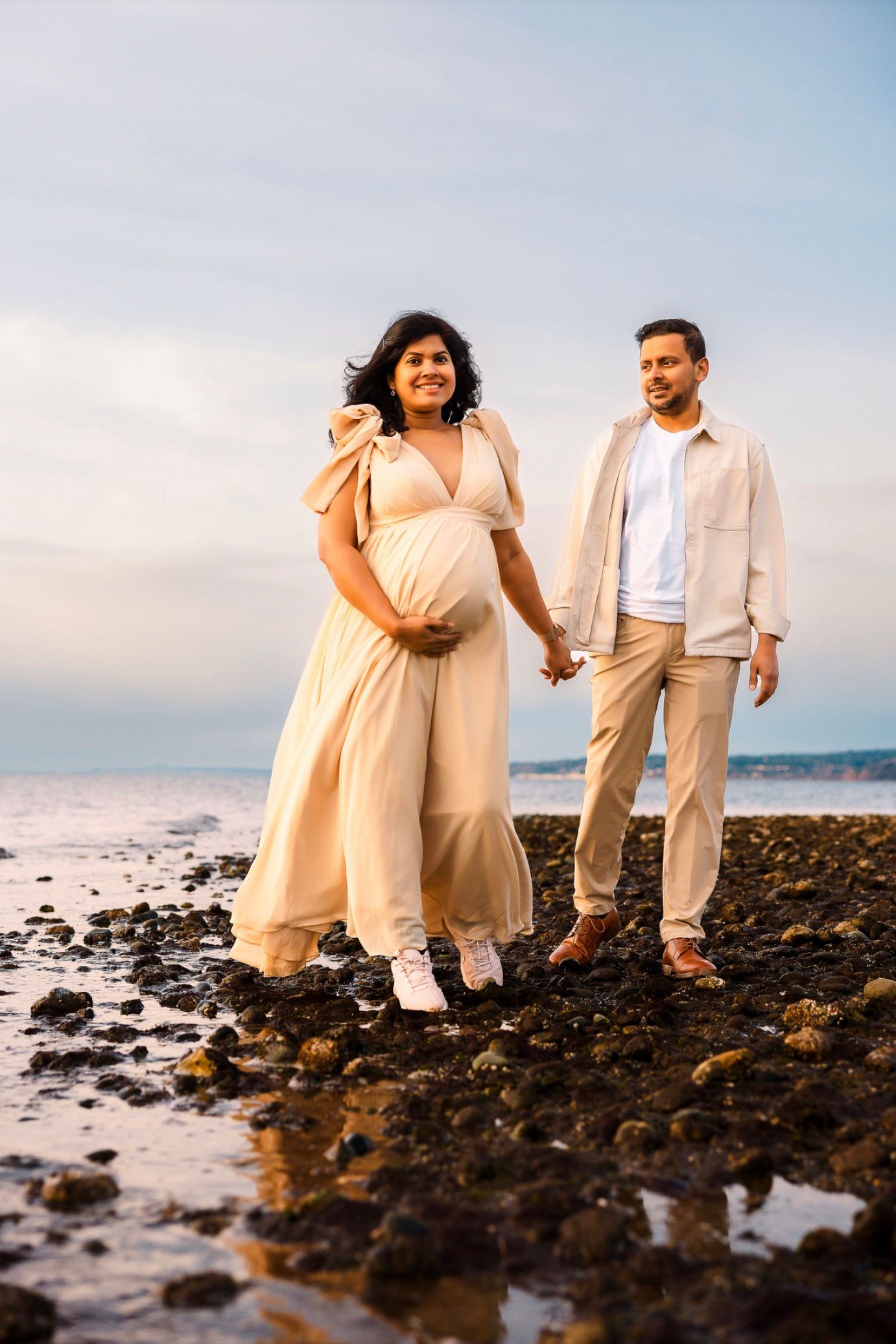 A couple walks along a rocky beach shoreline at sunset wearing white and peach colored outfits.