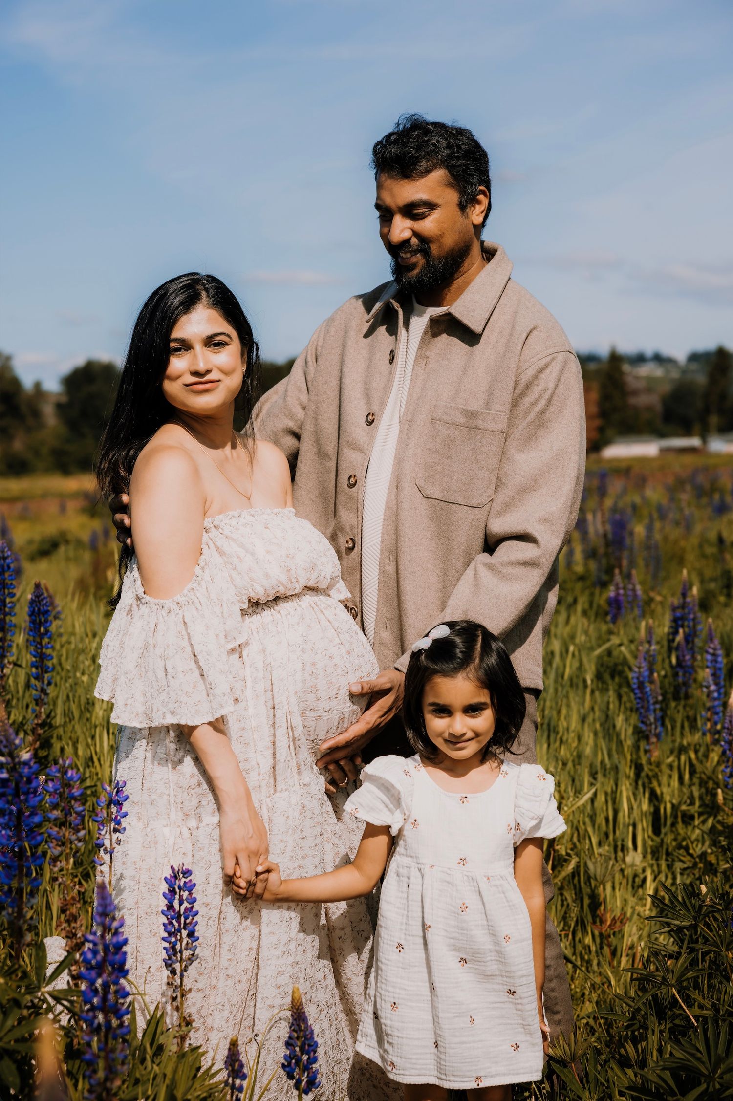 Pregnant woman in floral dress with husband and daughter posing in a lupine field for a maternity photo session.