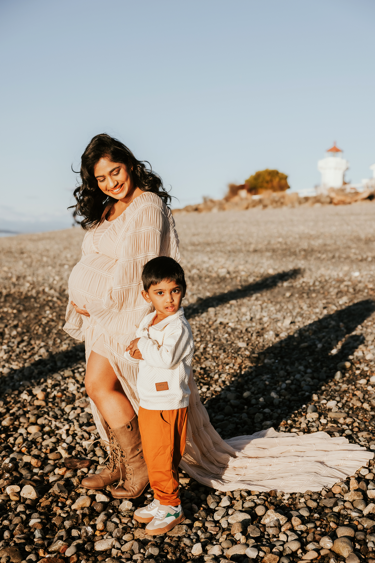 Mother and child sit together on a rocky beach at sunset with a lighthouse in the background.