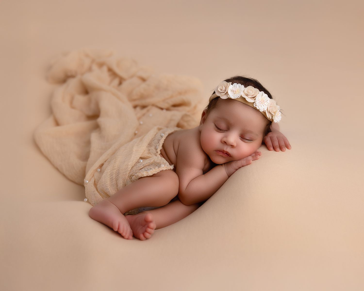 Sleeping newborn wearing a floral headband and cream wrap on beige backdrop during professional photo session.